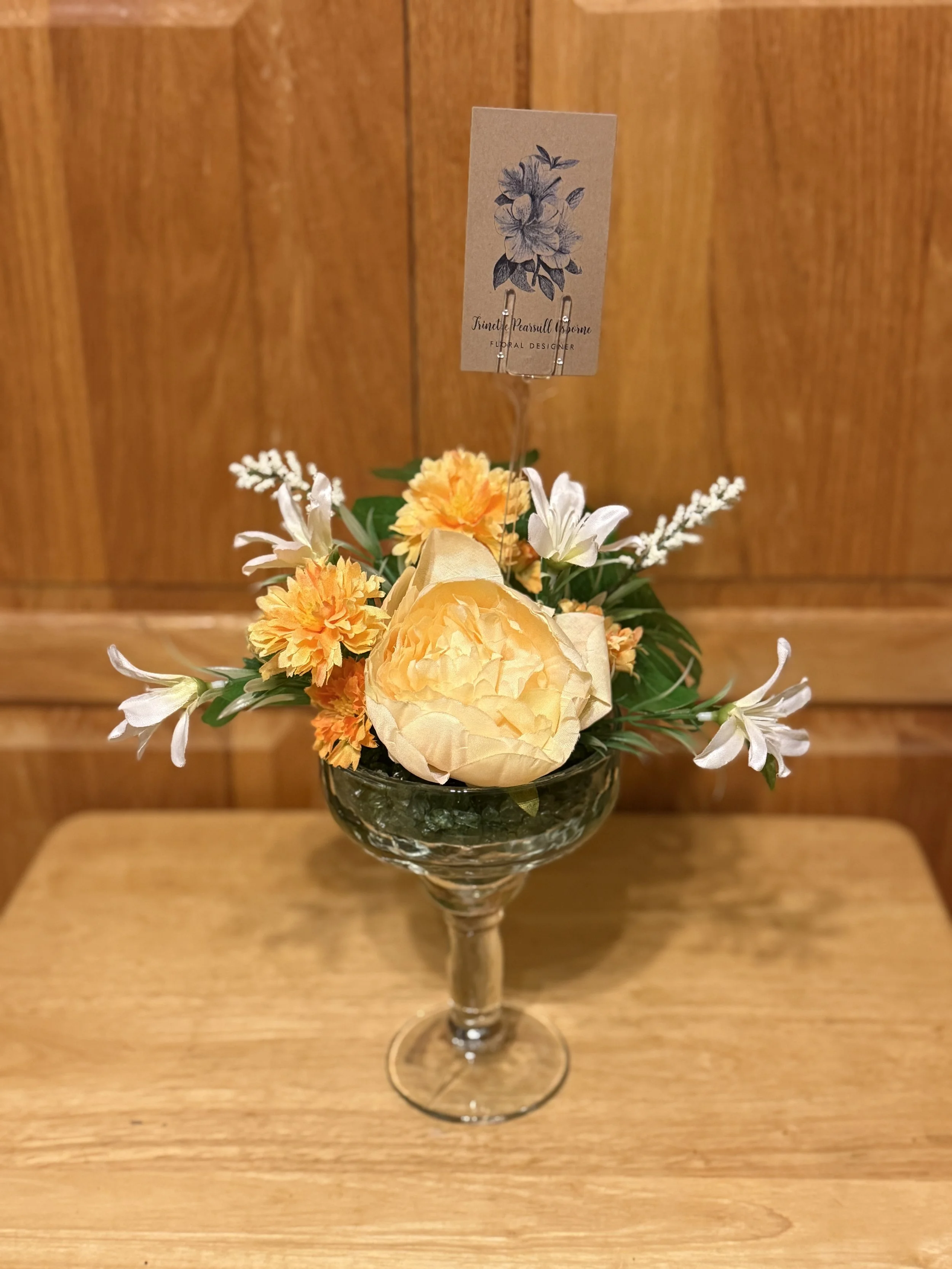 A floral arrangement in a glass goblet includes yellow roses, orange daisies, white lilies, and greenery with a card that has a blue floral illustration and the text 'Kenneth Pearsall Spence, Floral Designer'.