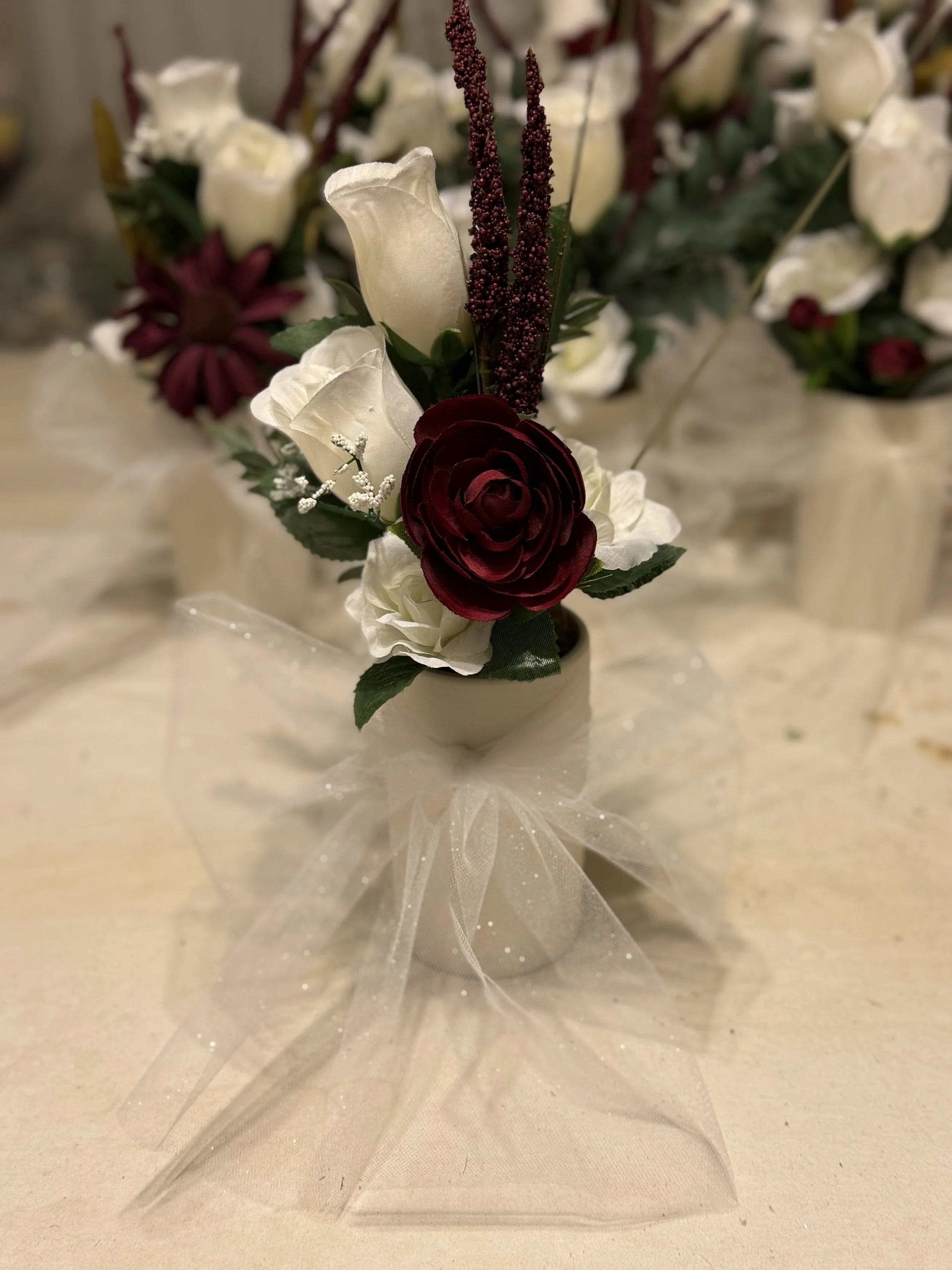 Small flower arrangement with white and dark red roses, green leaves, and purple accents, wrapped with white tulle on a table.