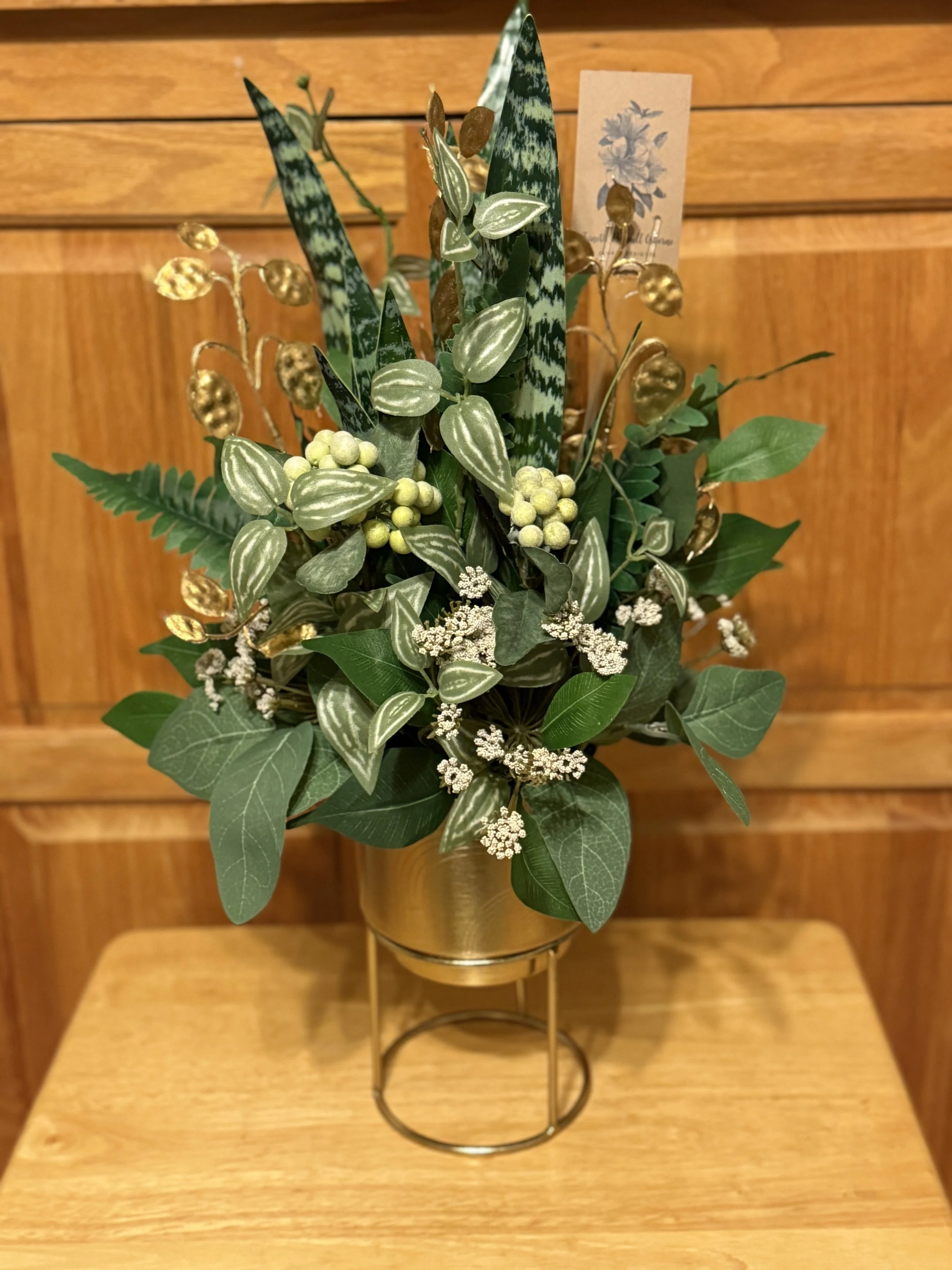 Artificial floral arrangement in a gold metal vase on a wooden table, with various green leaves, small white flowers, and golden leaves.