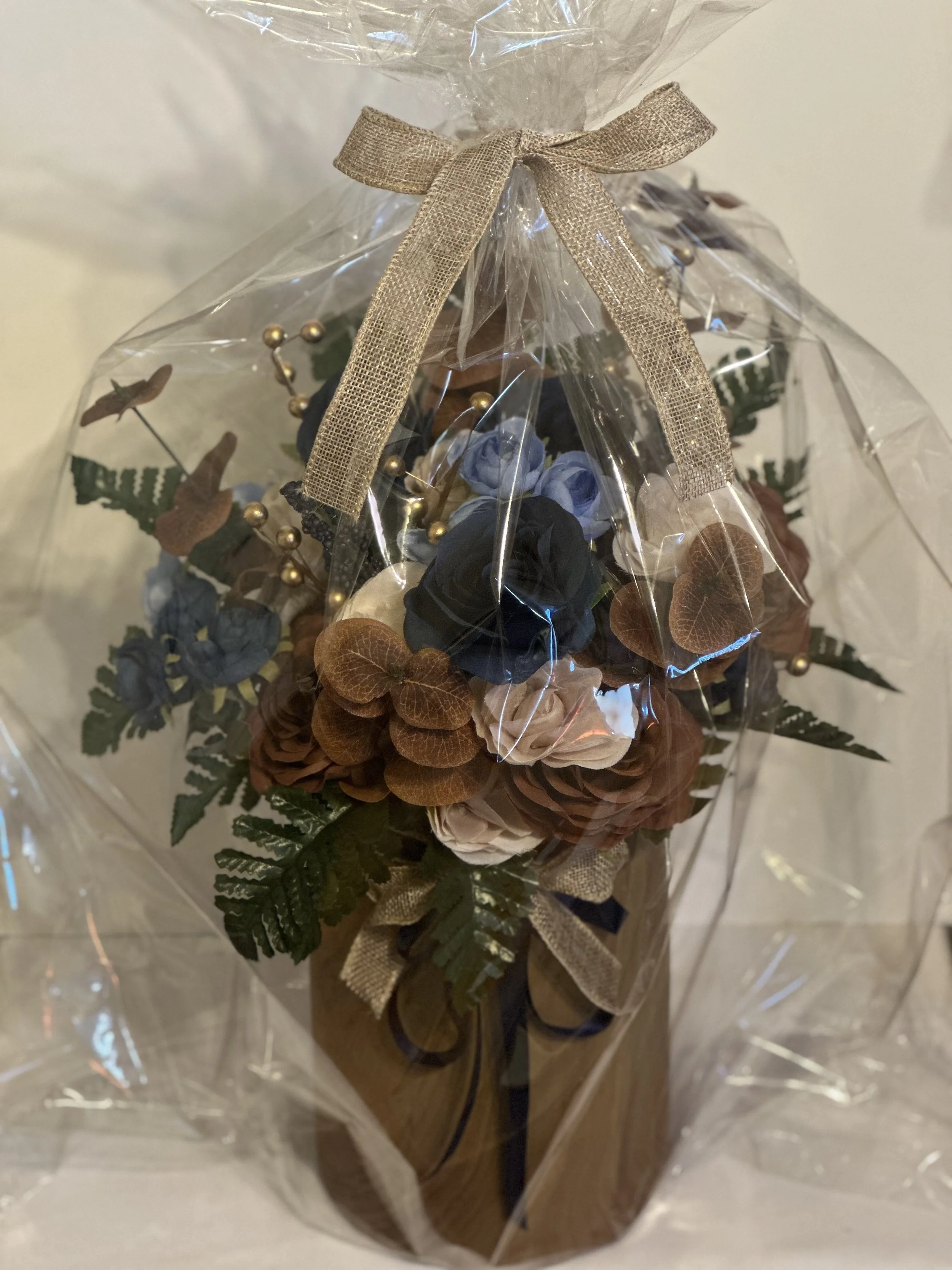 A decorative floral arrangement wrapped in clear plastic with a gold ribbon on top. The arrangement contains blue, brown, beige, and green artificial flowers and leaves, arranged in a wood grain vase.