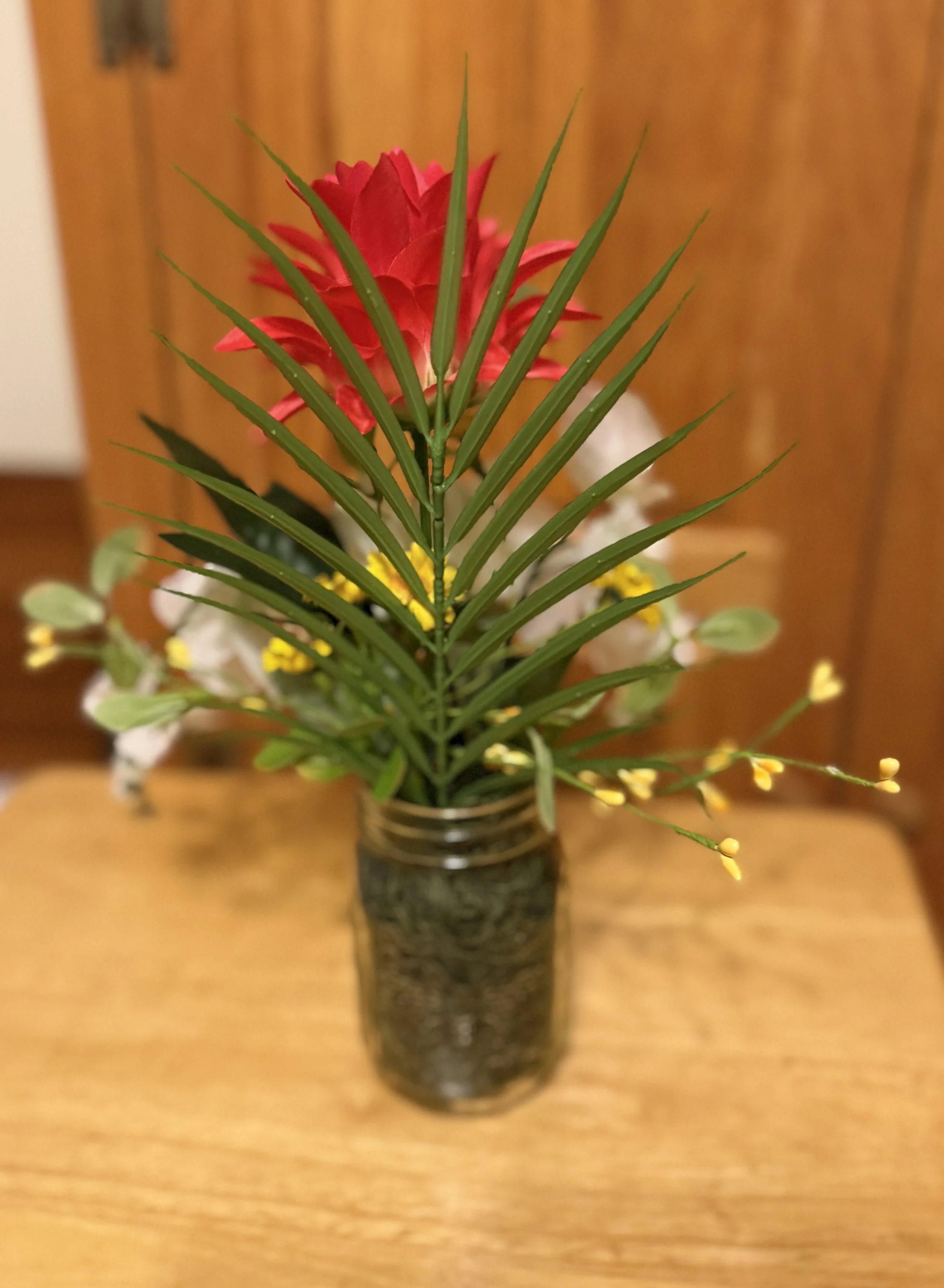 A small glass jar with floral artificial arrangement including green leaves, a red flower, and small yellow buds.