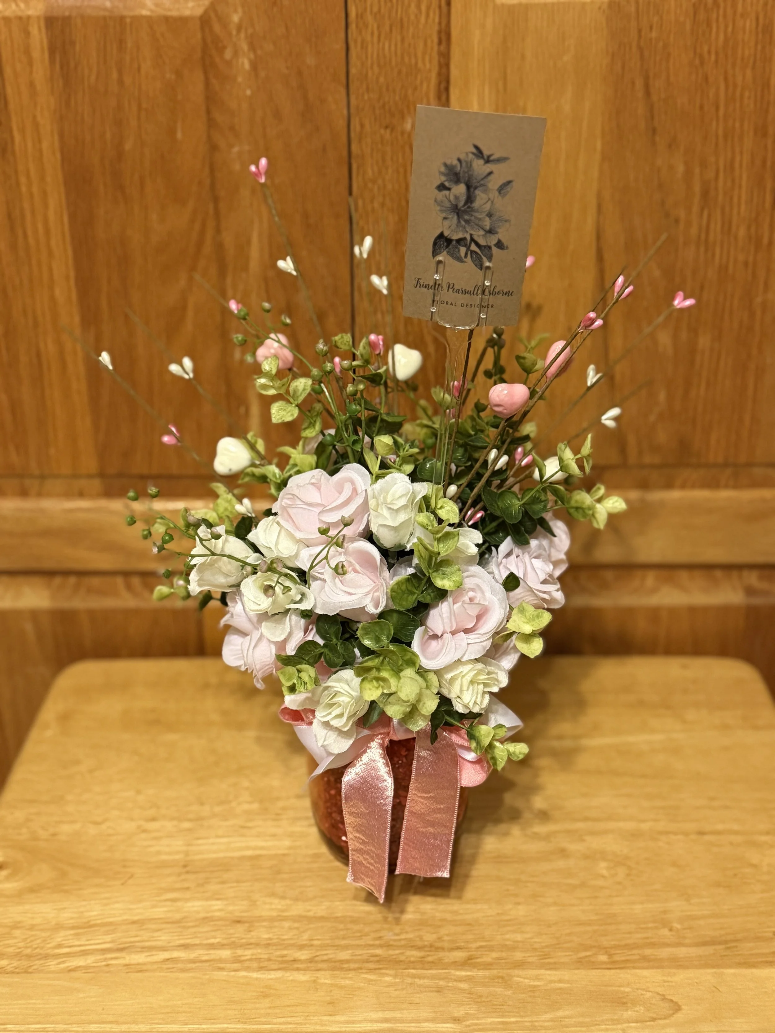 A flower arrangement with white and pink blossoms in a vase, decorated with a pink satin ribbon, placed on a wooden surface against a wooden wall background.