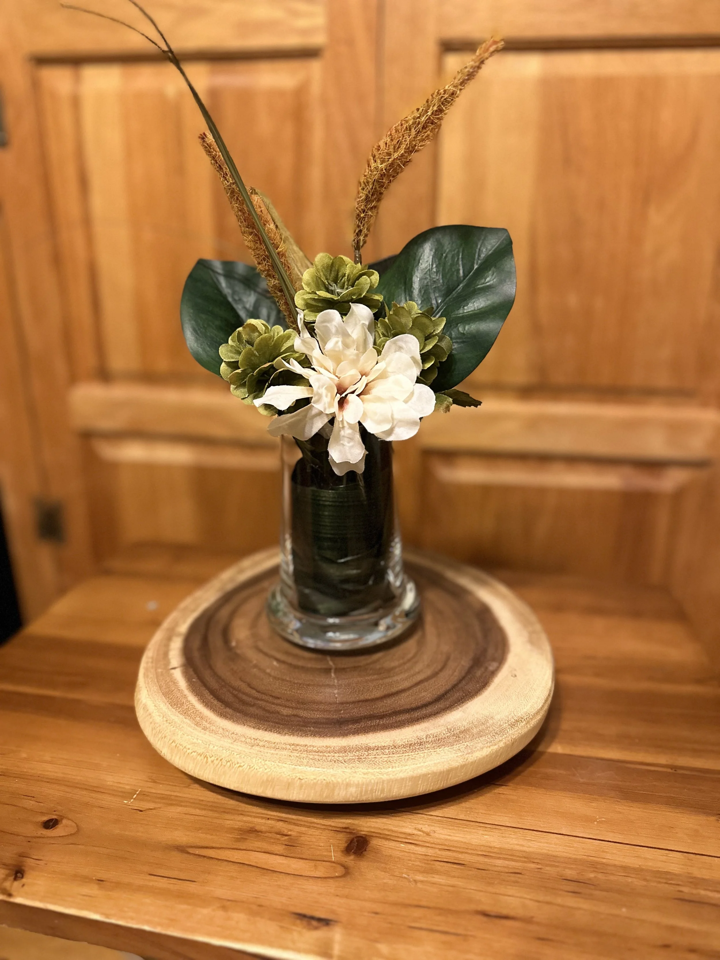 A floral arrangement in a clear glass vase with white and green flowers, large green leaves, and ornamental grasses, placed on a round wooden tray on a wooden table.