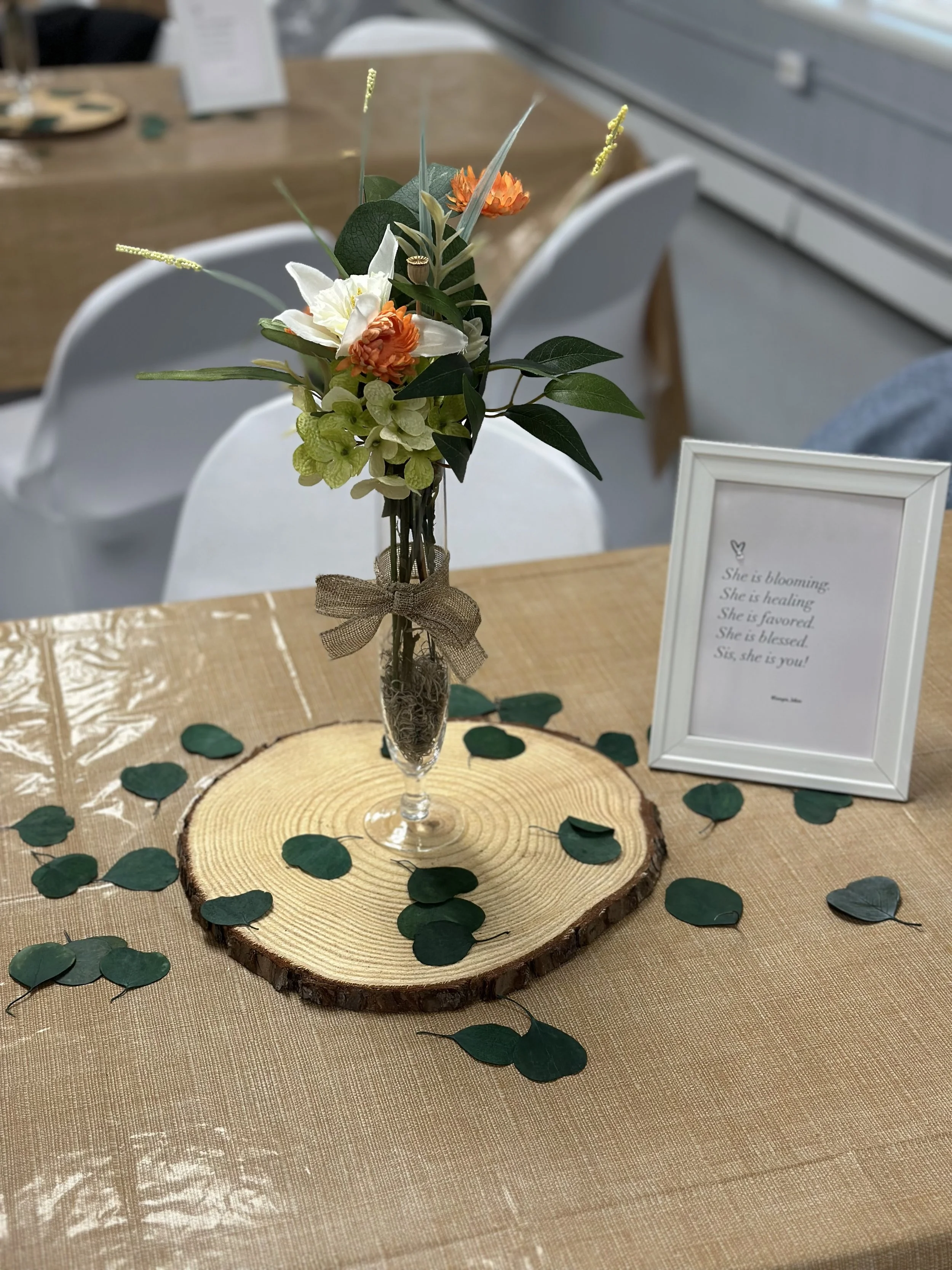 A floral centerpiece in a clear glass vase with a burlap ribbon, placed on a round wooden slab on a beige tablecloth with scattered green leaves. A white-framed sign with a poem stands beside it.