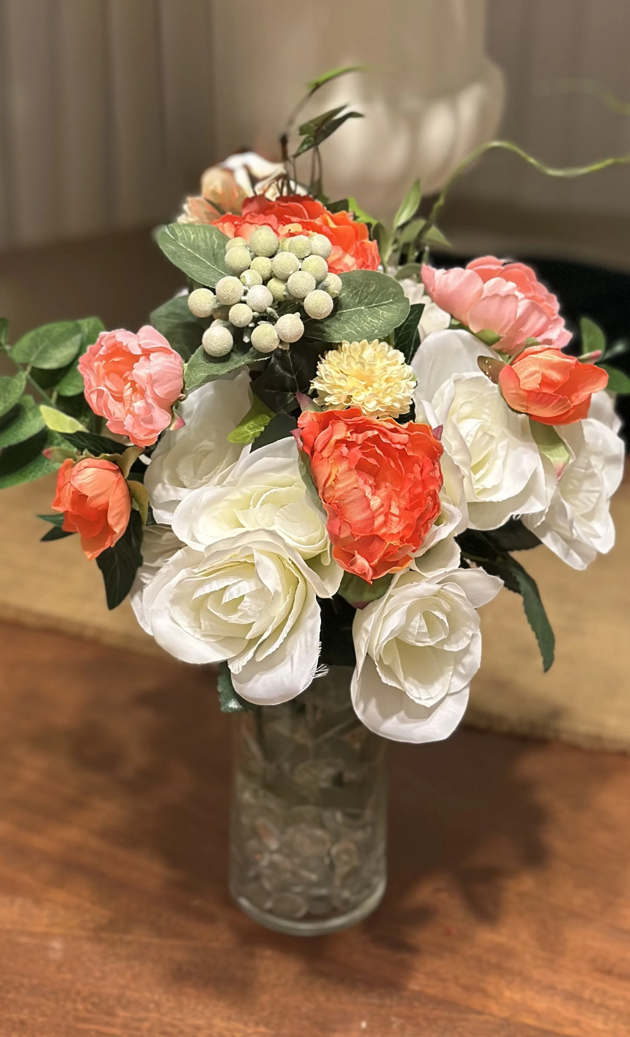 A bouquet of artificial flowers in a glass vase on a wooden table, consisting of white roses, pink and orange peonies, green foliage, and white berries.