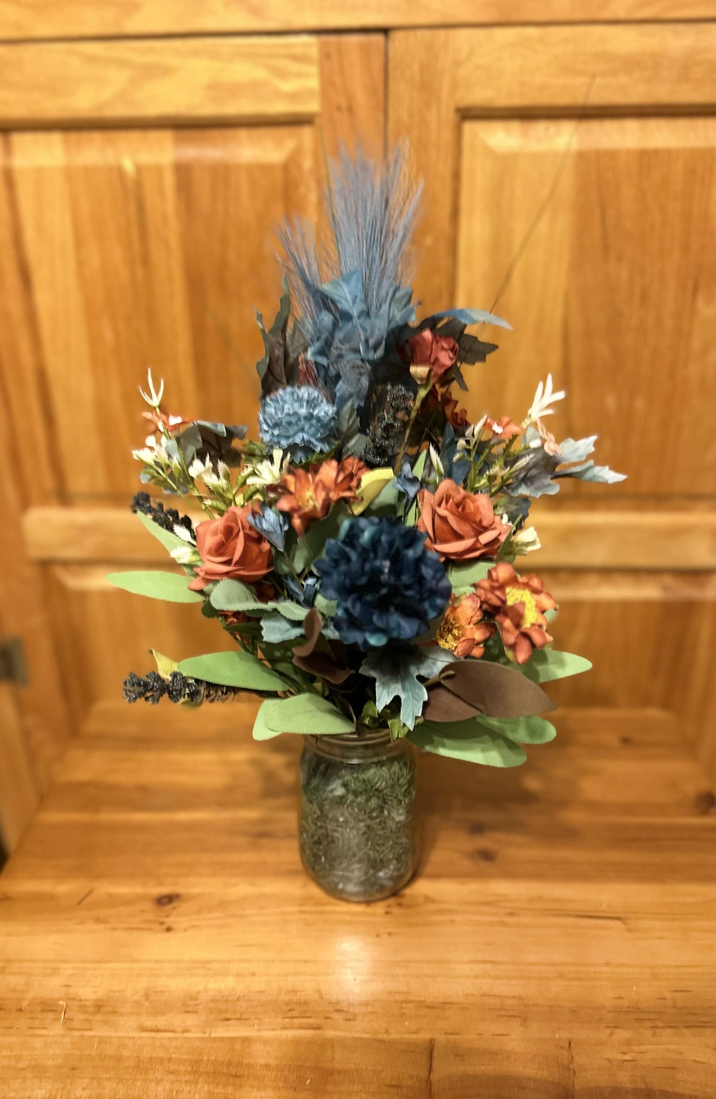 Artificial flower arrangement with orange, blue, black, and beige flowers and green leaves in a glass jar, set against a wooden background.
