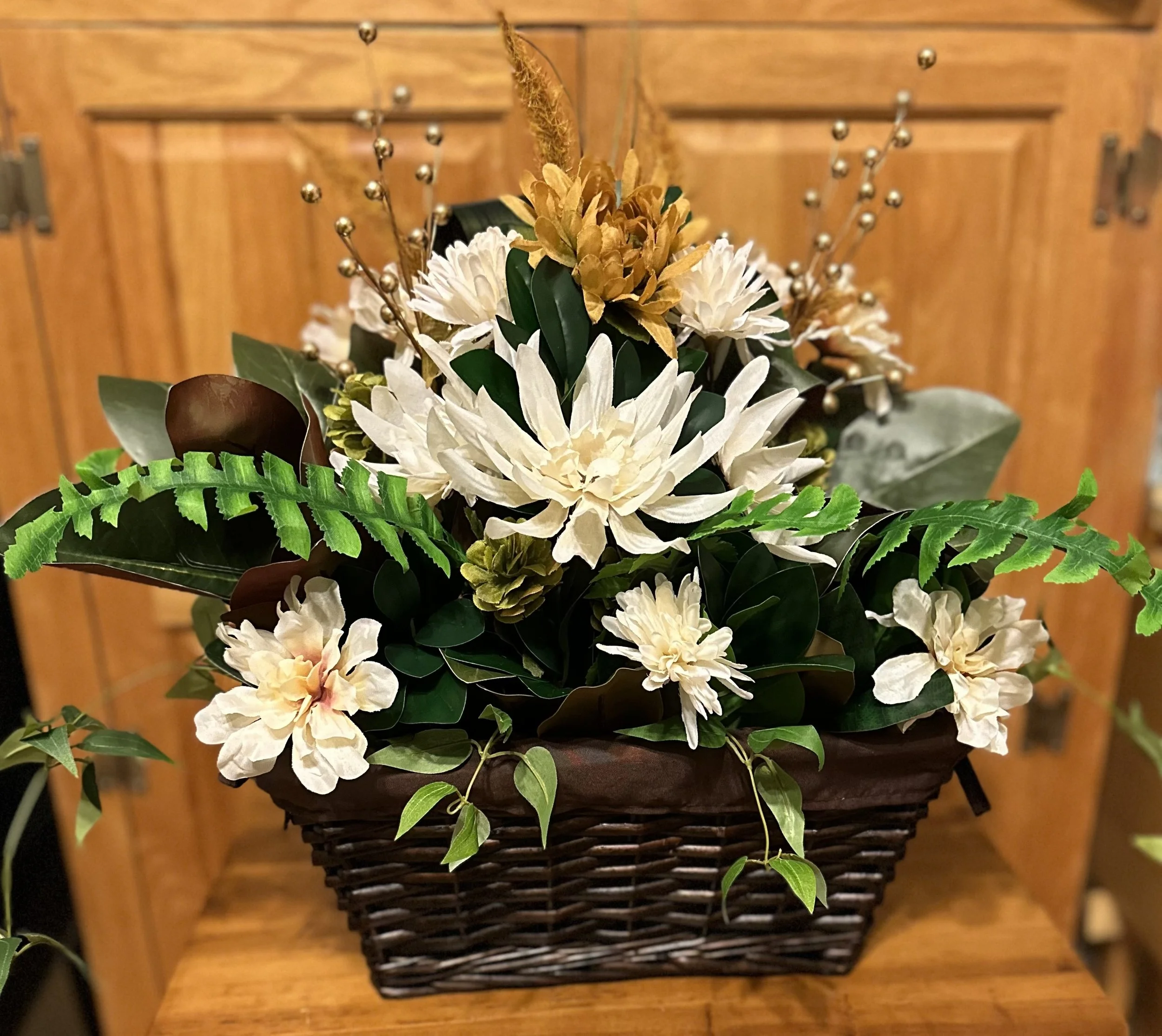 A flower arrangement in a basket with white and beige flowers, green leaves, and gold accents, placed on a wooden surface with a wooden cabinet in the background.