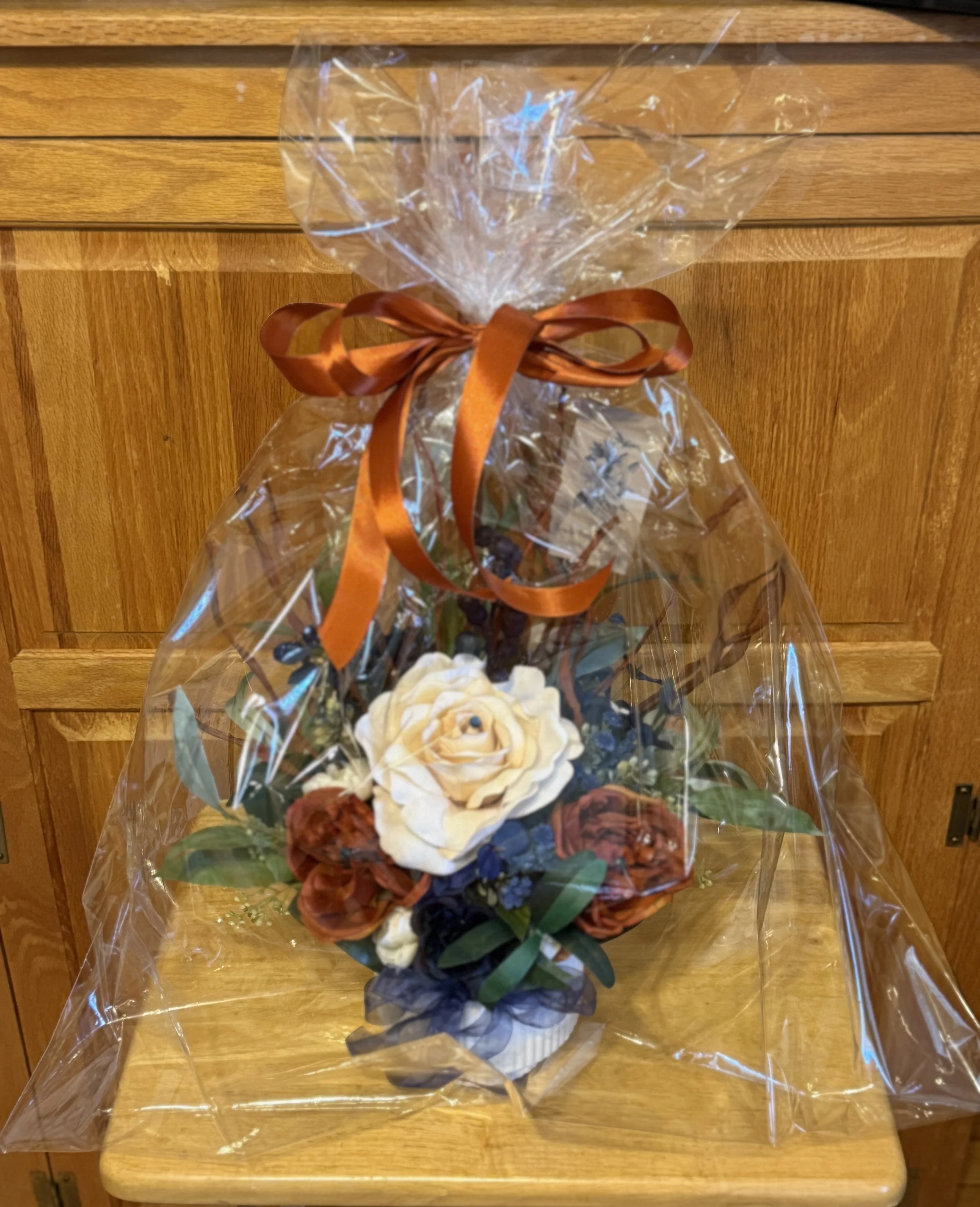 An arrangement consisting of silk peonies and artificial flowers including white, rust, and blue flowers, wrapped in clear plastic with a ribbon,.