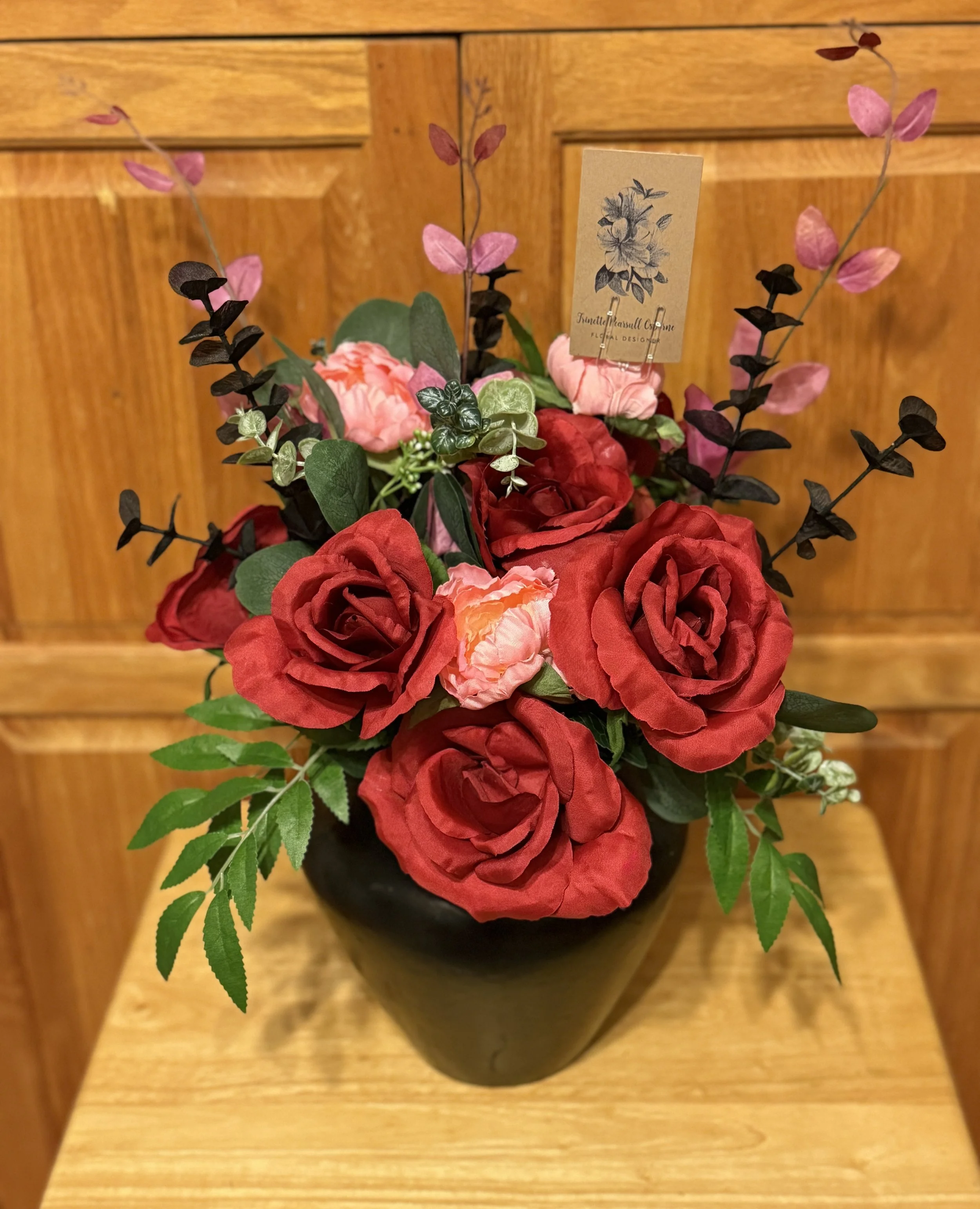 A vase of artificial red and pink roses with green leaves and purple accents on a wooden surface against a wooden paneled background.