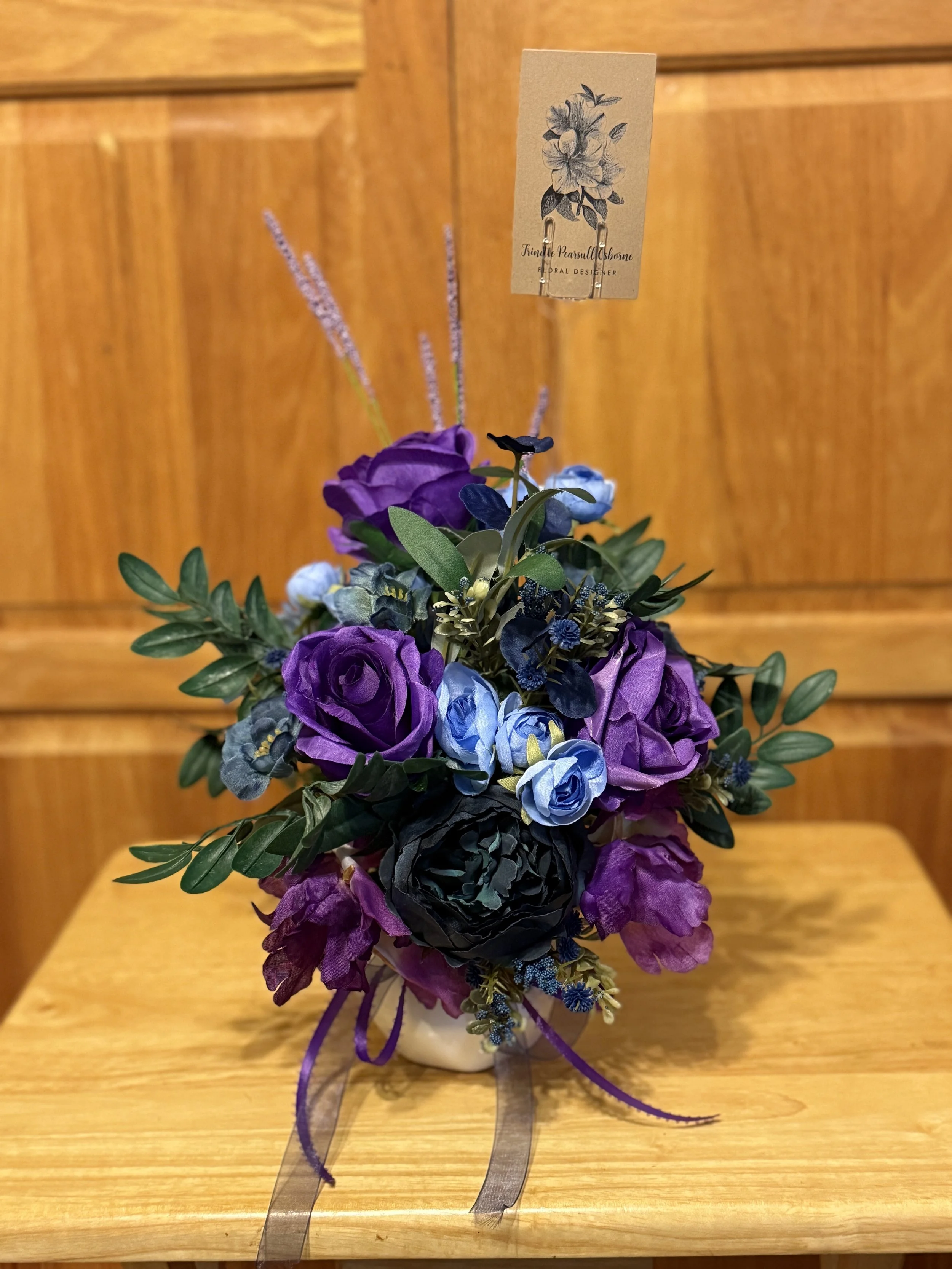 A small floral arrangement with purple, blue, artificial flowers in a white vase, decorated with ribbons.