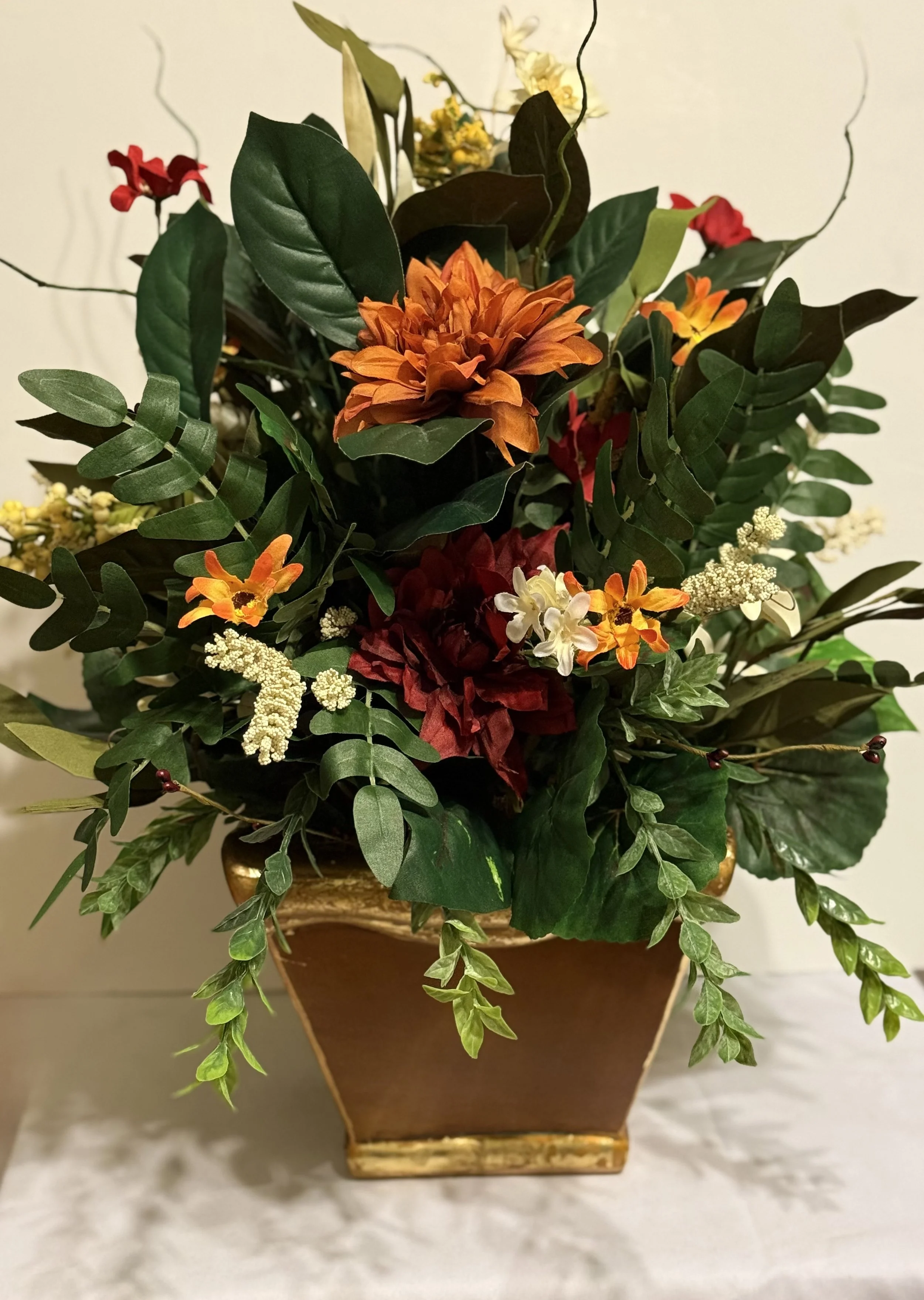 A decorative floral arrangement in a gold-colored square pot featuring orange, red, white, and yellow flowers with green leafy foliage.
