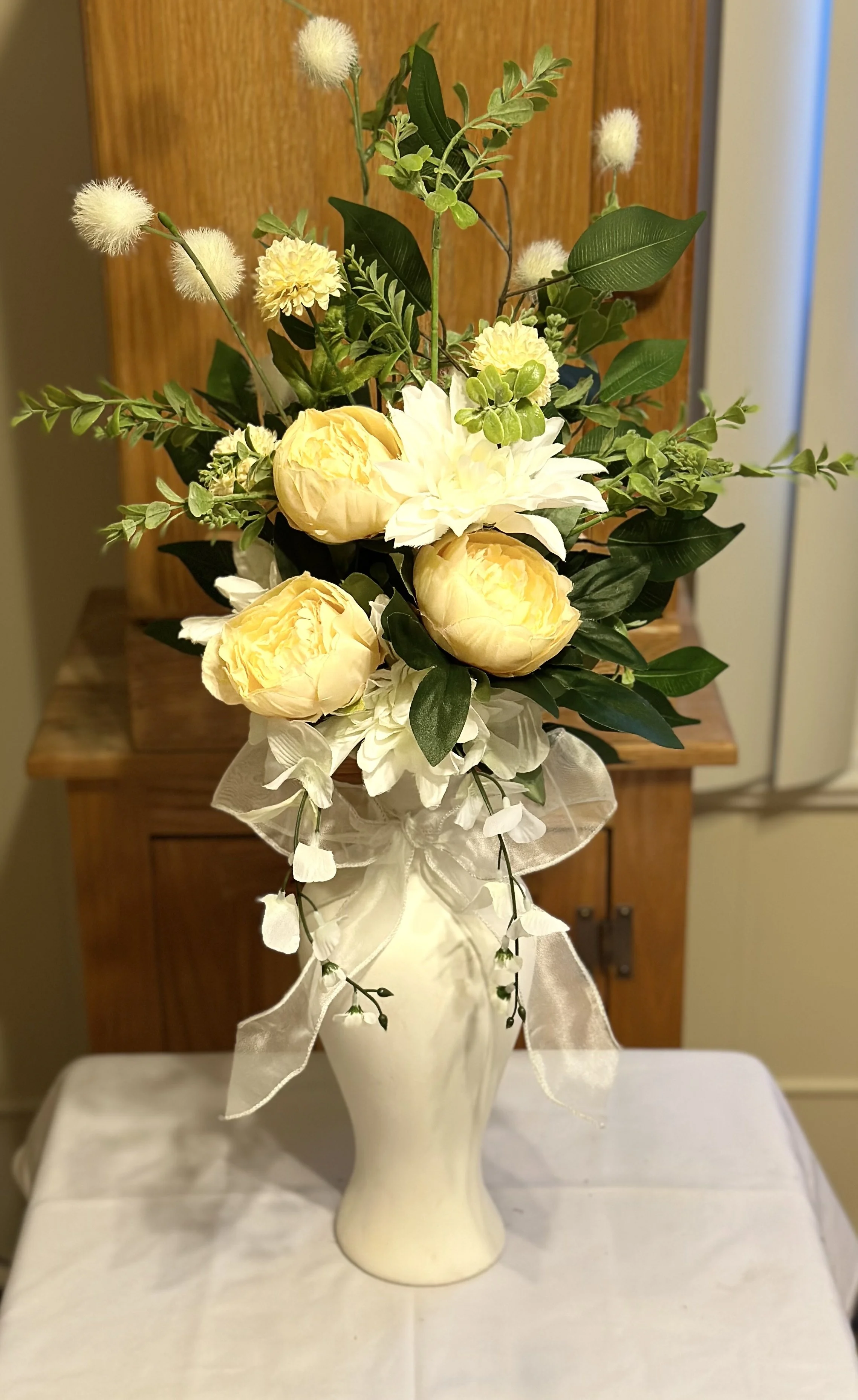 A floral arrangement in a cream-colored vase, featuring yellow peonies, white dahlias, green leaves, and small white fluffy flowers, decorated with a sheer ribbon tied around the neck of the vase.