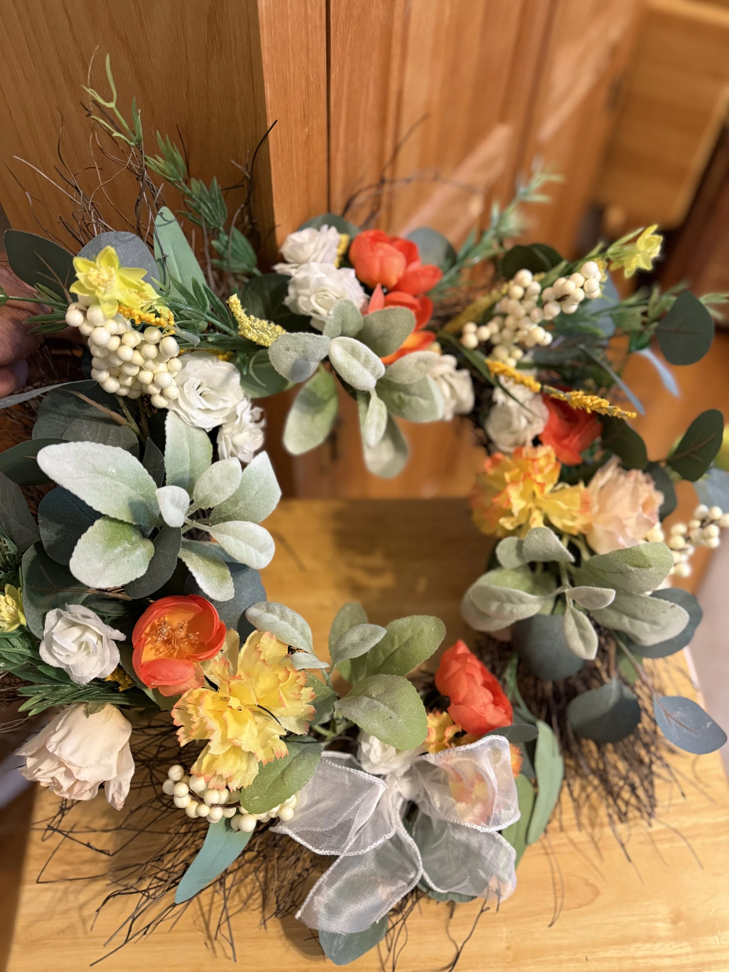 A decorative floral wreath with white, yellow, orange, and red flowers, green leaves, berries, and a white sheer ribbon on a wooden surface.