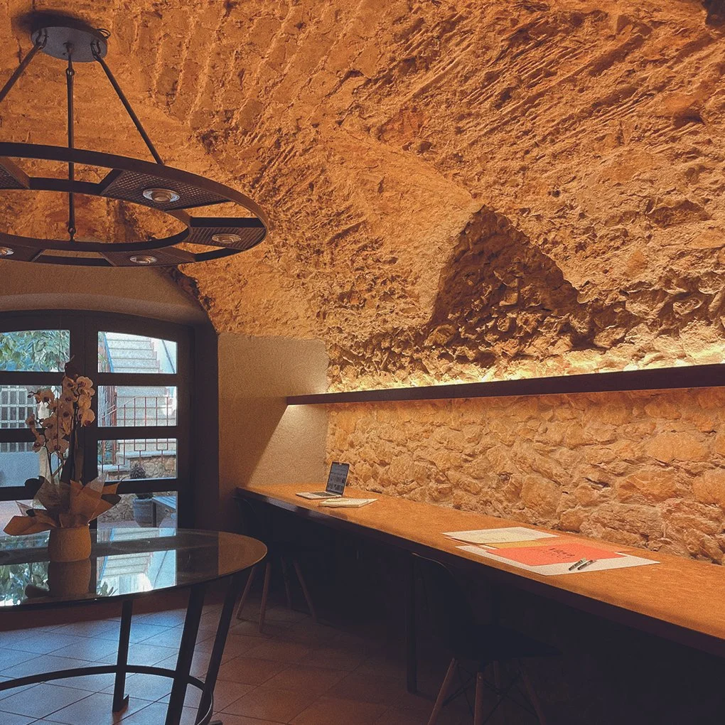 Interior de un restaurante con paredes de piedra, una mesa con flores, una ventana y una lámpara moderna en el techo.