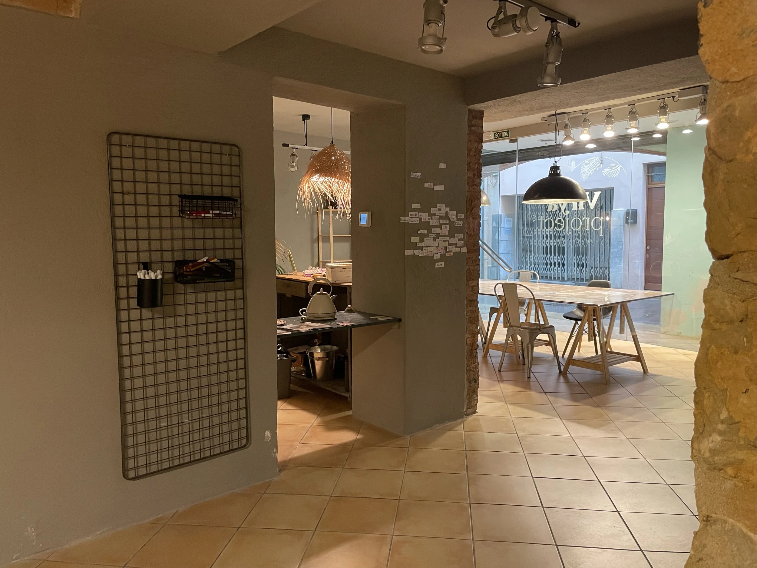 Interiores de un café con mesas de madera y sillas, ventanas grandes con vista a la calle, lámparas modernas y decoración sencilla.