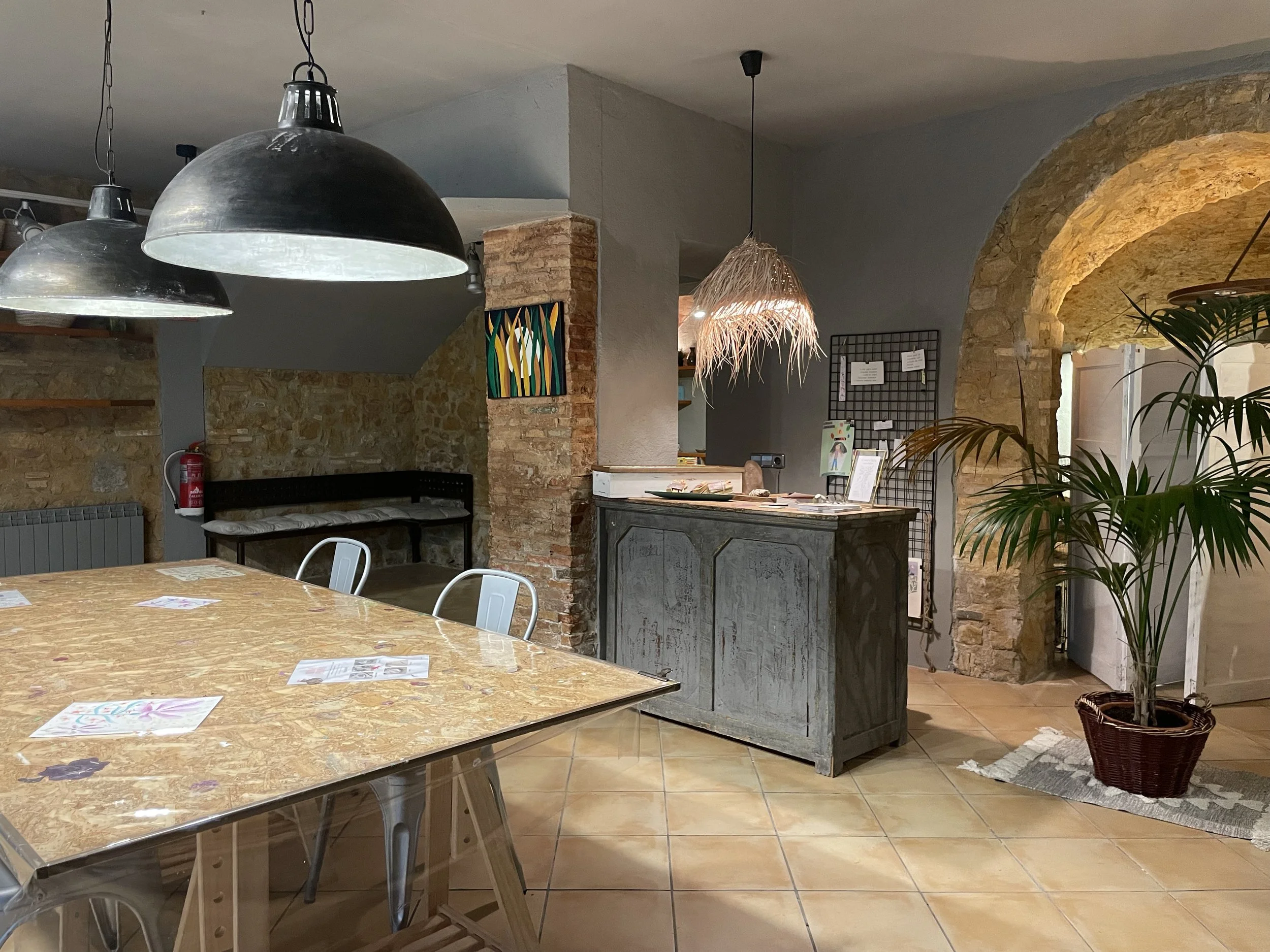 Interior de un restaurante con mesa de madera, sillas metálicas, lámparas colgantes, una planta en una maceta de mimbre y una barra de atención con decoración rústica con piedra y ladrillo.