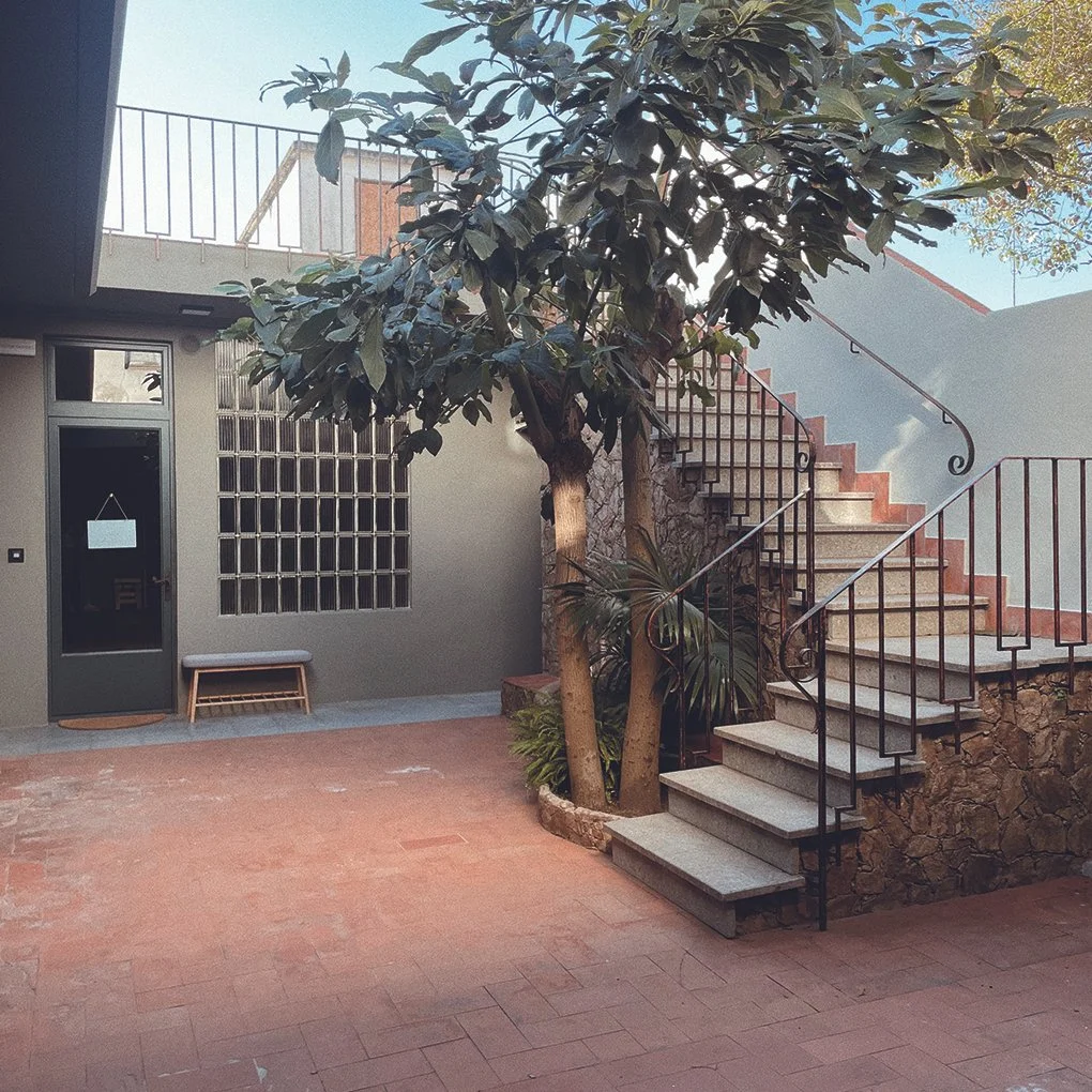 Vista de un patio con escaleras de piedra y barandillas de hierro, con un árbol en el centro y una entrada con una puerta de cristal y un banco.
