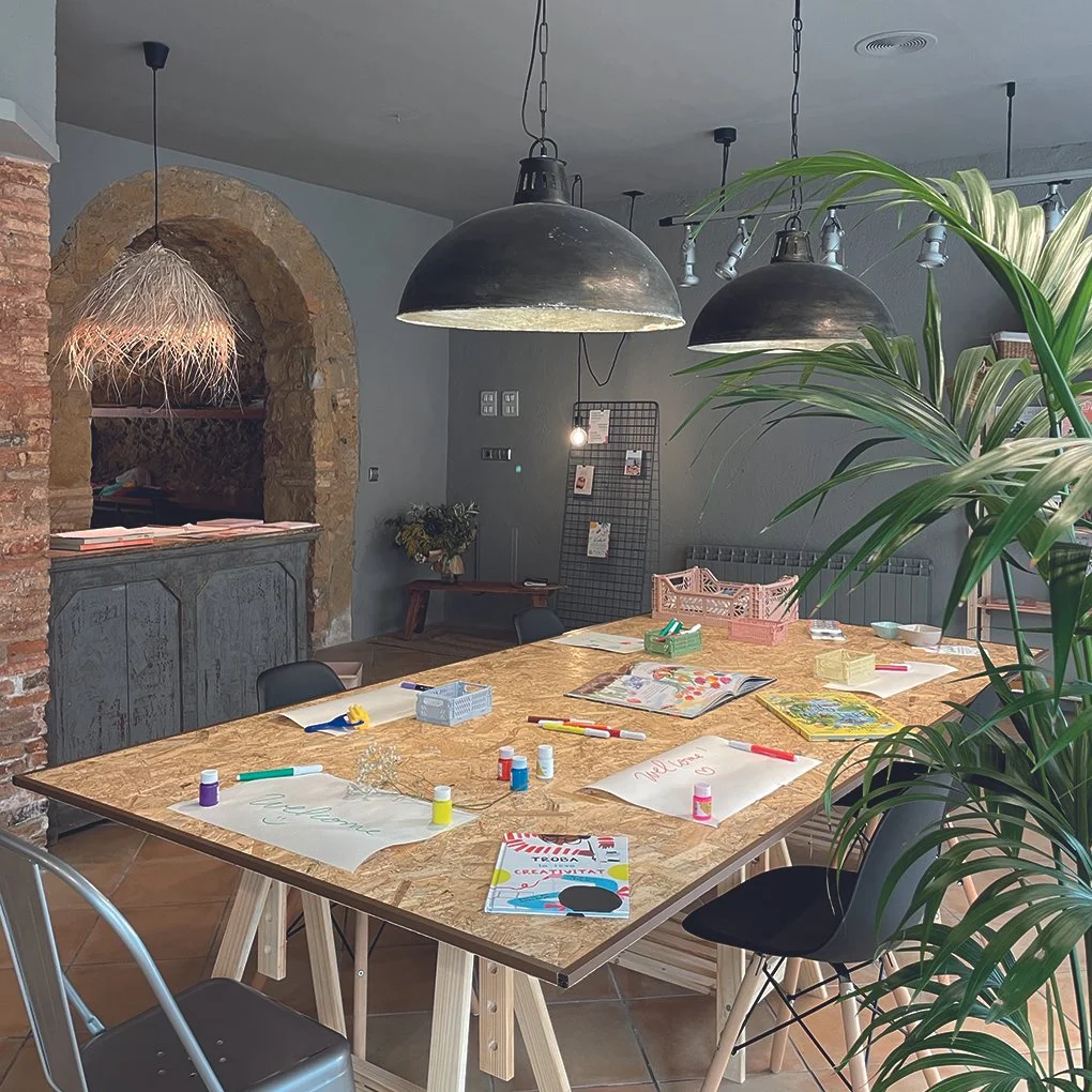 Una mesa de madera con materiales para arte y manualidades, rodeada de sillas negras, en un espacio decorado con plantas, lámparas negras colgantes y una pared de ladrillos a la vista. En el fondo, hay una pared gris con una malla decorativa y una me