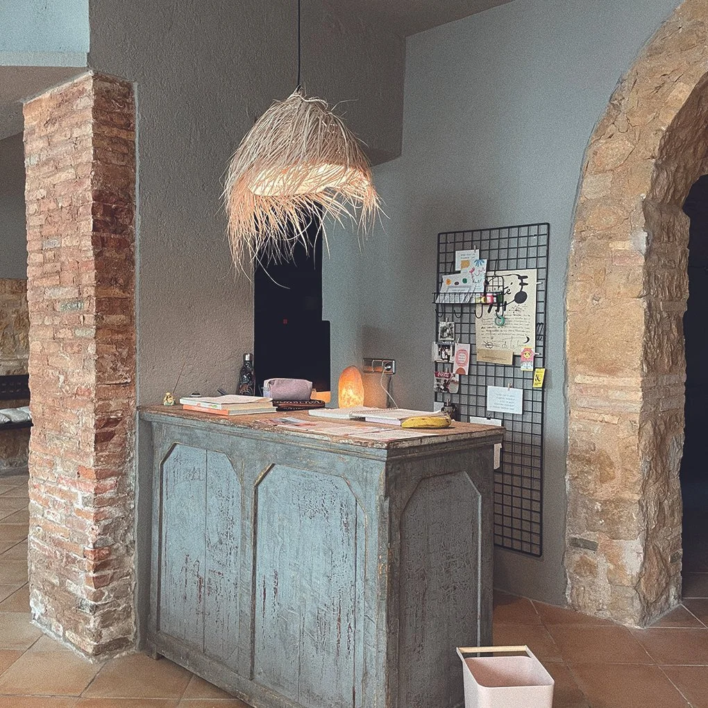 Mostrador de madera pintada en tonos azules envejecidos, con objetos decorativos y libros sobre su superficie, en un ambiente con paredes de ladrillo y piedra, y una lámpara de fibra natural colgando del techo.