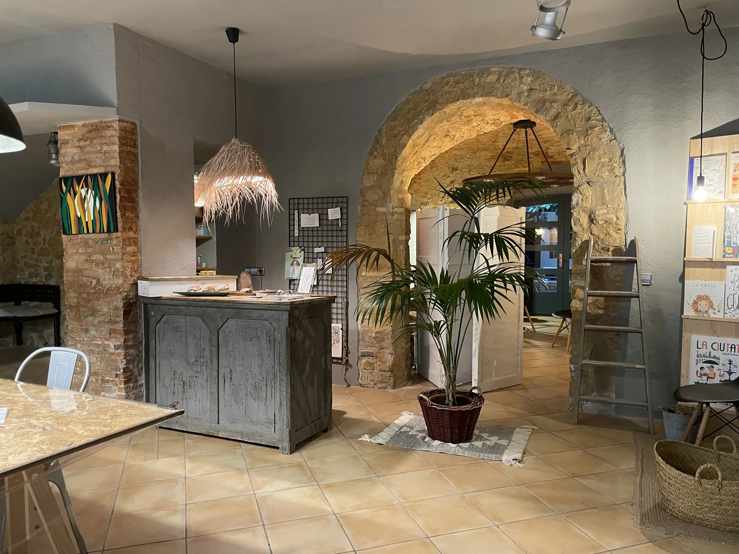 Interior de un cafe o librería con paredes de ladrillo, una planta en una maceta de mimbre, un mostrador de madera pintada de gris desgastado, estanterías con libros y una pared con un arcos de piedra.