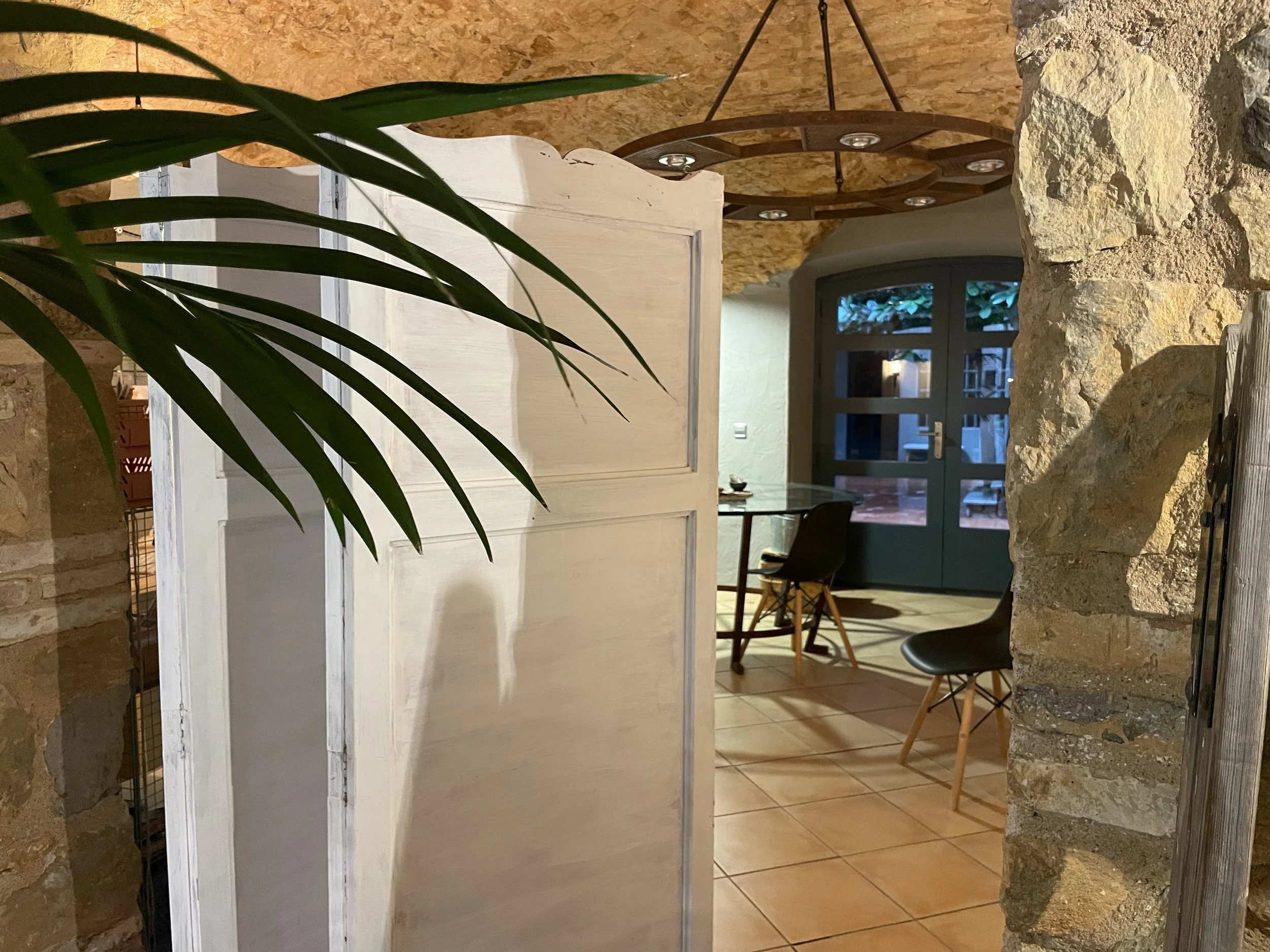 Vista de un comedor con paredes de piedra, una mesa redonda de vidrio, sillas negras de madera, una puerta doble de madera y vidrio, y una lámpara circular decorativa en el techo, con plantas visibles en el fondo.