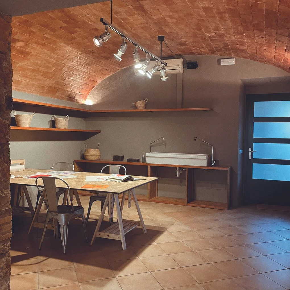 Sala de cocina con mesa de madera, sillas metálicas, estanterías con cestas y utensilios, fregadero y puerta de entrada de vidrio mate en la esquina.