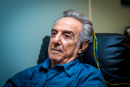 Elderly man with gray hair and a serious expression sitting in a medical chair with electrodes attached, in a clinical setting.