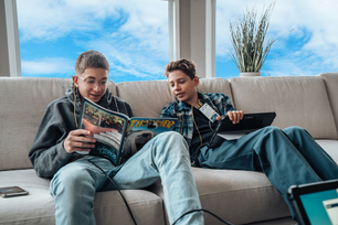 Two boys sitting on a beige couch reading comic books and using a tablet in a bright living room with large windows and a plant behind them.