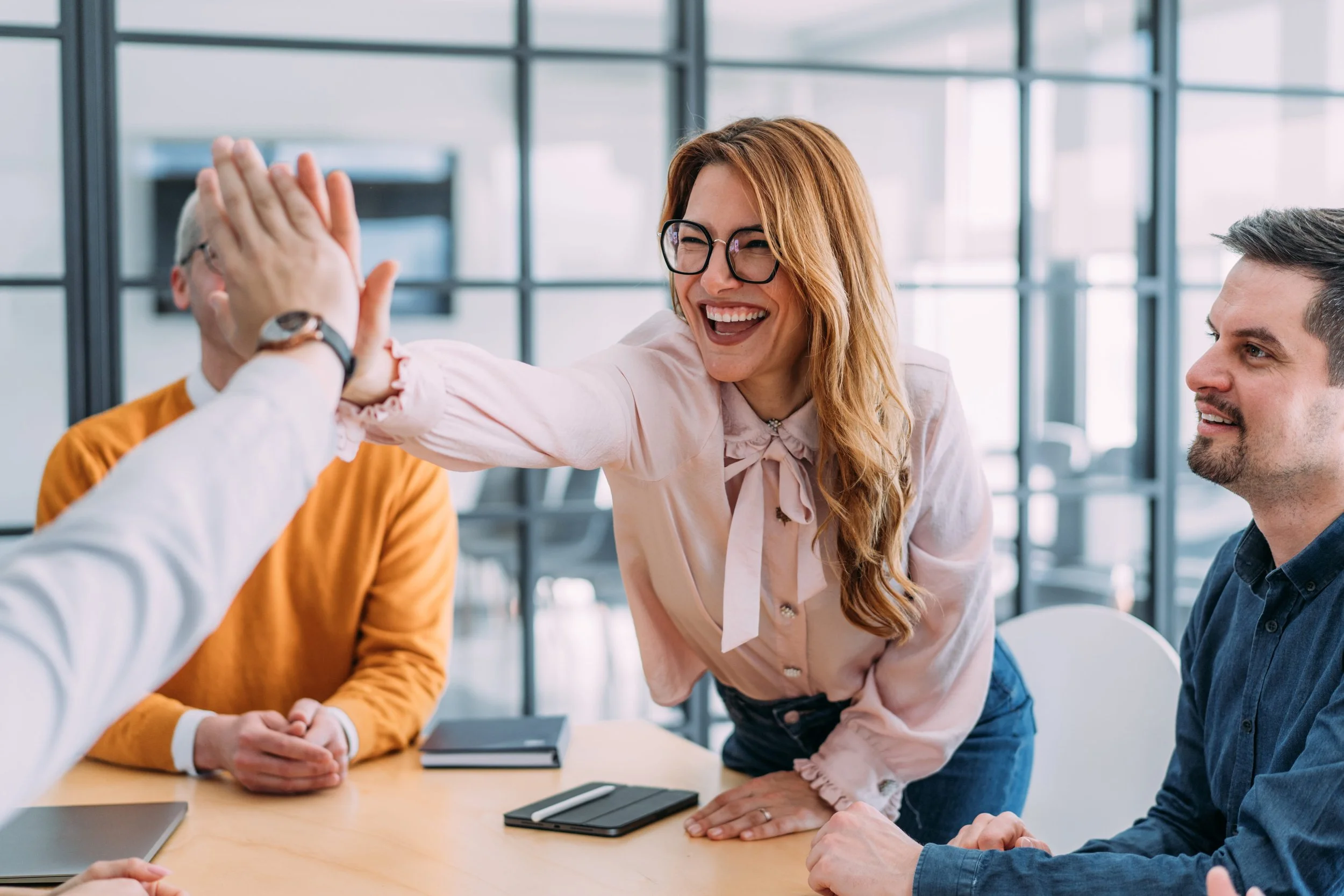 A woman with glasses smiling and giving a high five to a man during a meeting in an office.