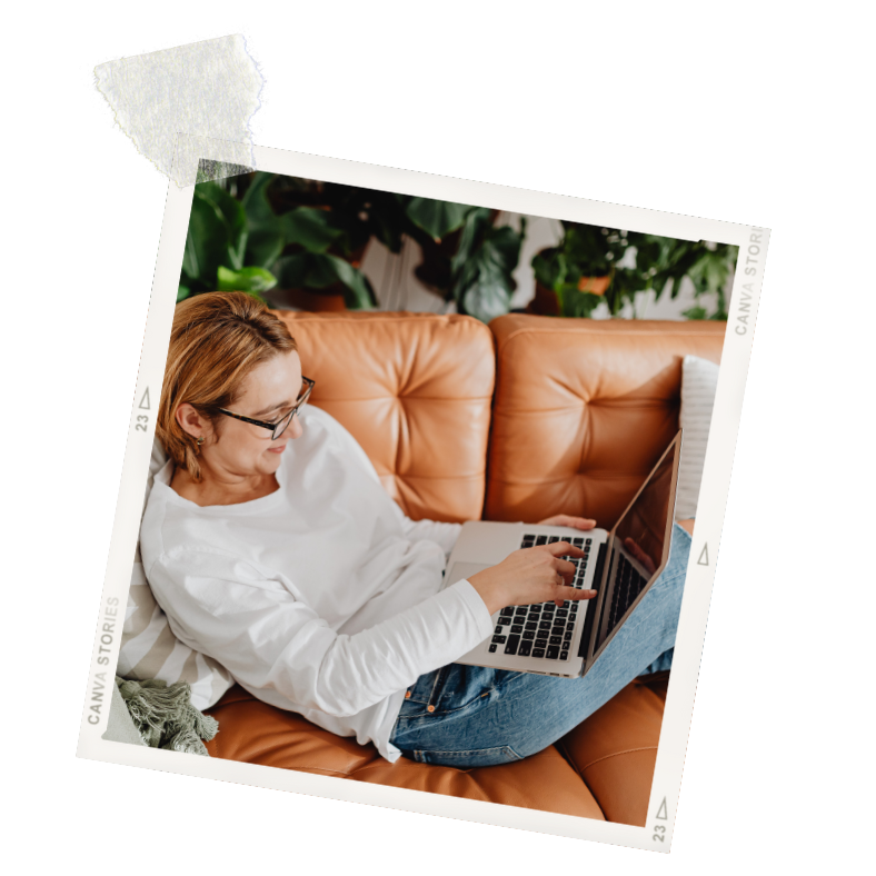 A woman with glasses, wearing a white shirt and jeans, sitting on a tan leather couch, using a laptop.