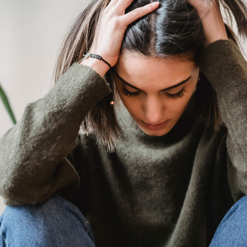 A woman with short brown hair and long eyelashes, wearing a green sweater, is sitting with her head down and hands on her head.
