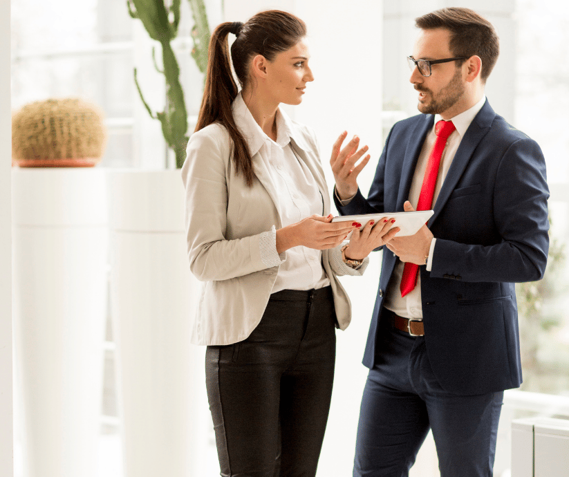 A woman and a man in business attire having a discussion in an office, with the woman holding a tablet.