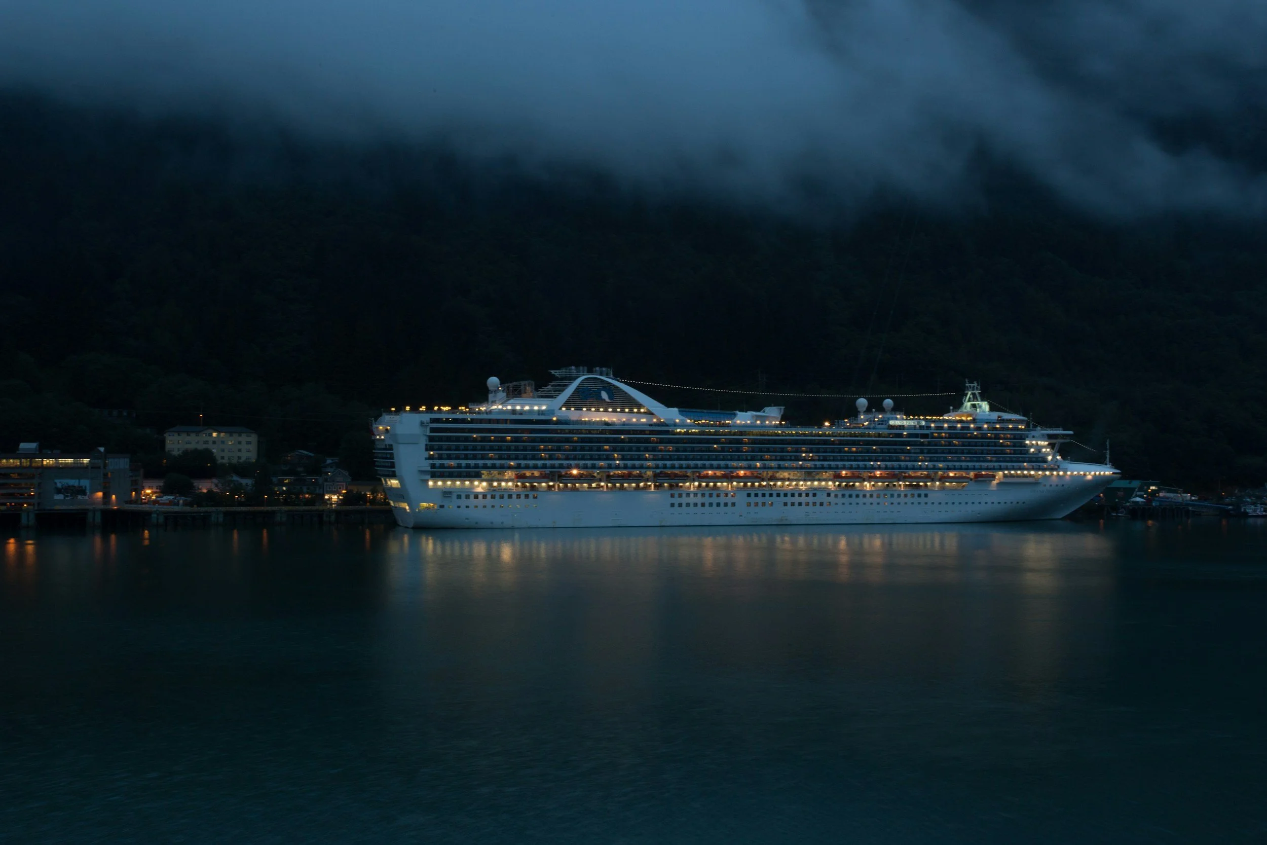 A large cruise ship docked at a port at night, with city lights and dark hills in the background.