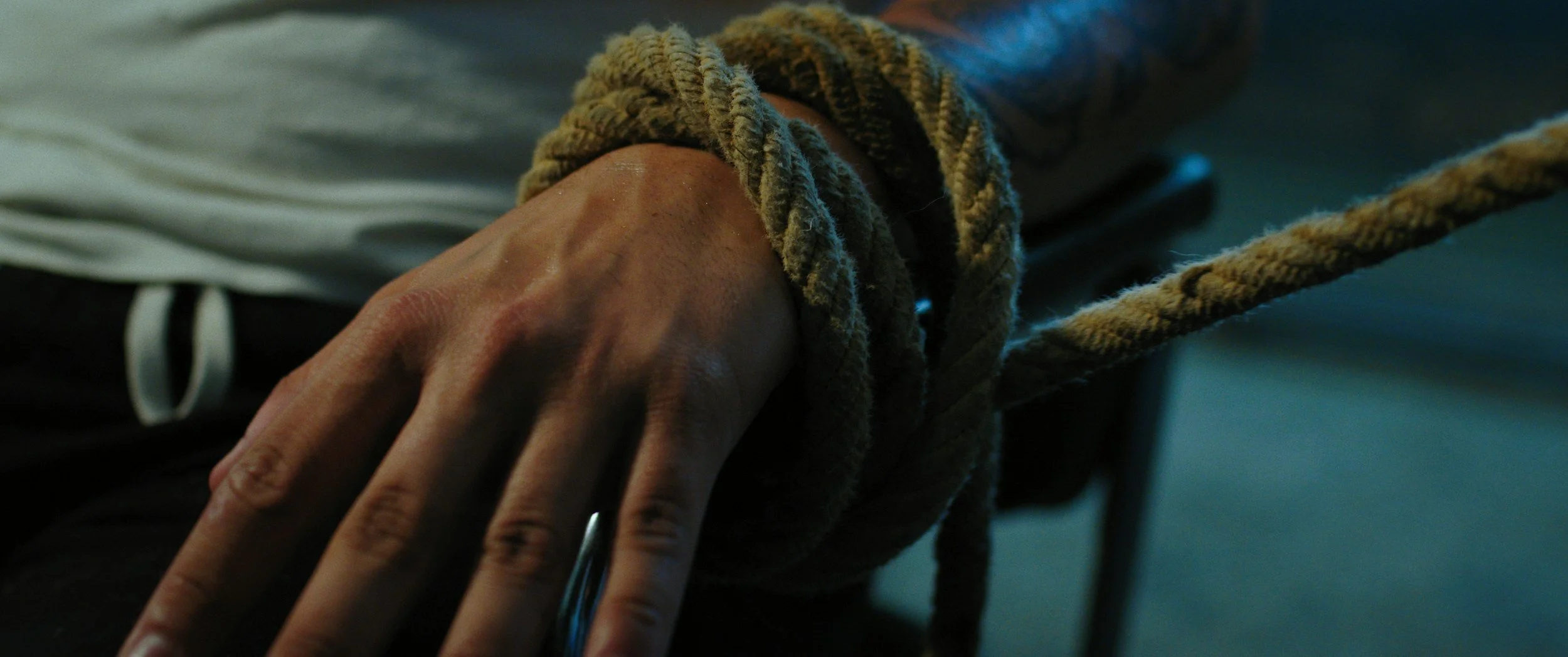 A hand with tied rope around the wrist, resting on a surface, with part of a white shirt and black pants visible in the background.