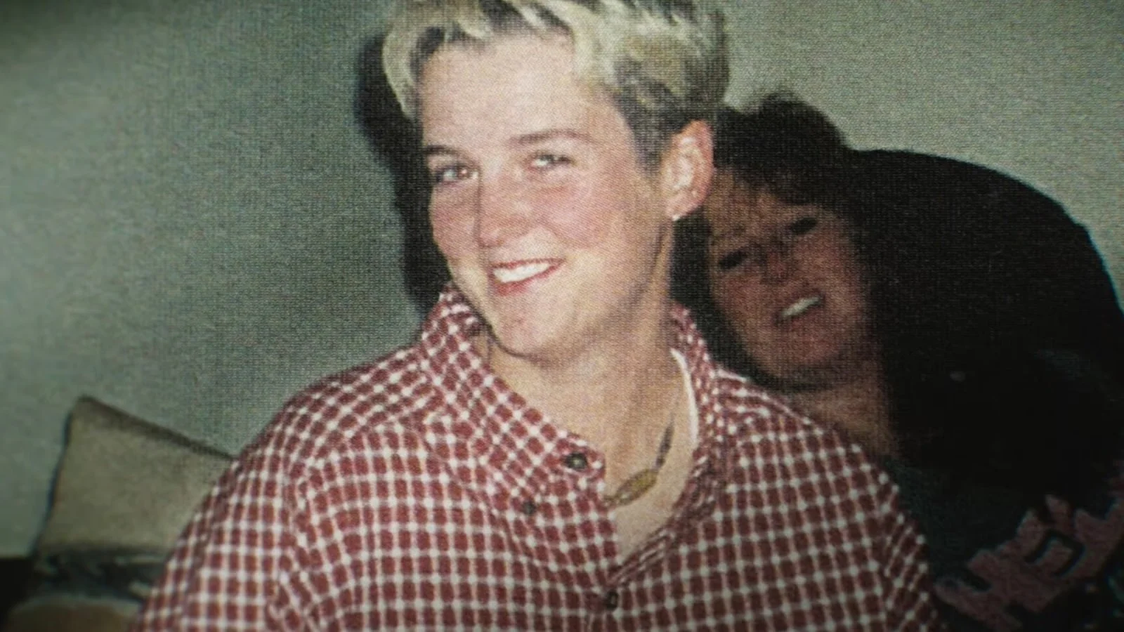 A woman with short blonde hair smiling, wearing a red and white checkered shirt and a necklace. In the background, another woman with glasses appears to be making a funny face.