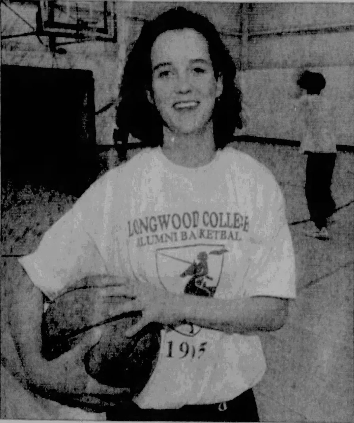 A young woman with shoulder-length dark hair, smiling, holding a basketball, wearing a "Longwood College Alumni Basketball" T-shirt with the year 1975 printed on it, standing inside a gymnasium.