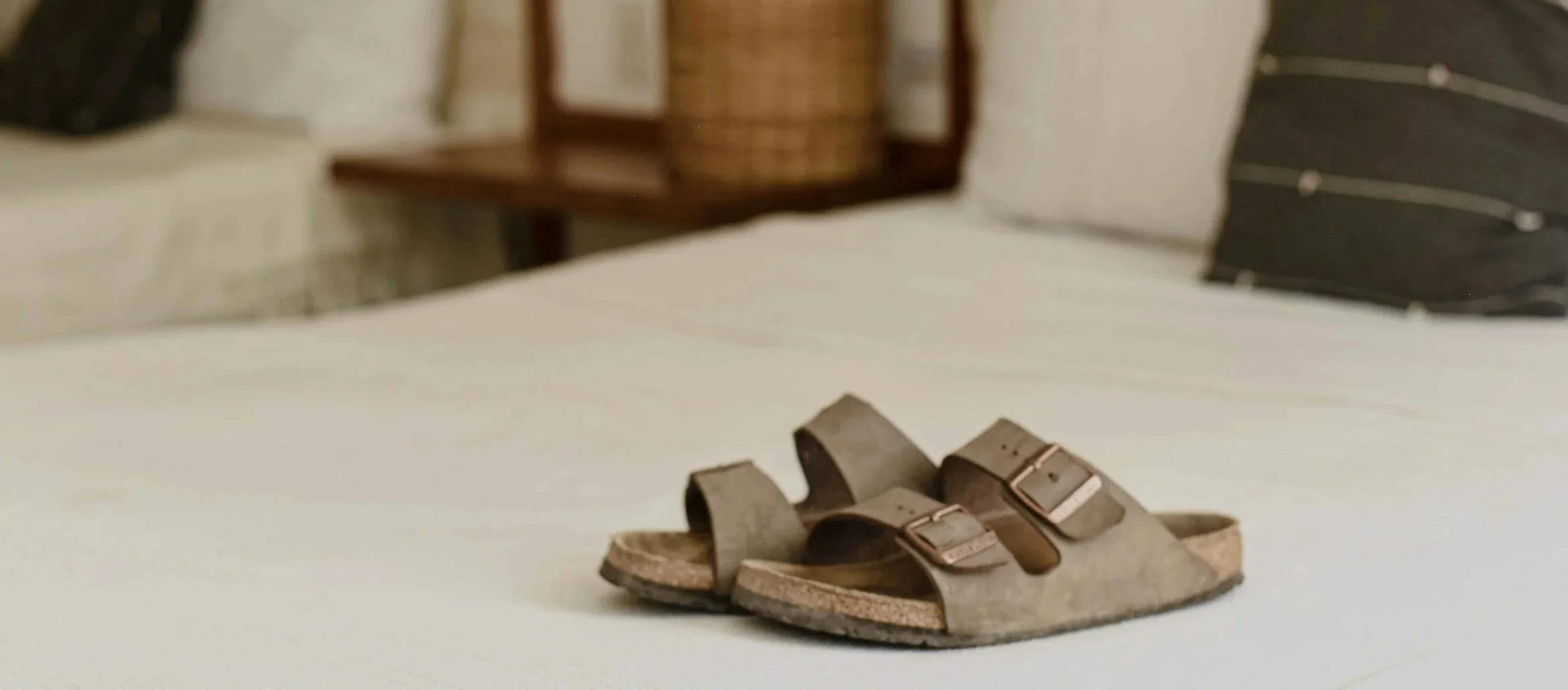 A pair of brown sandals with two buckles on a white surface, with a bed and other furniture in the background.