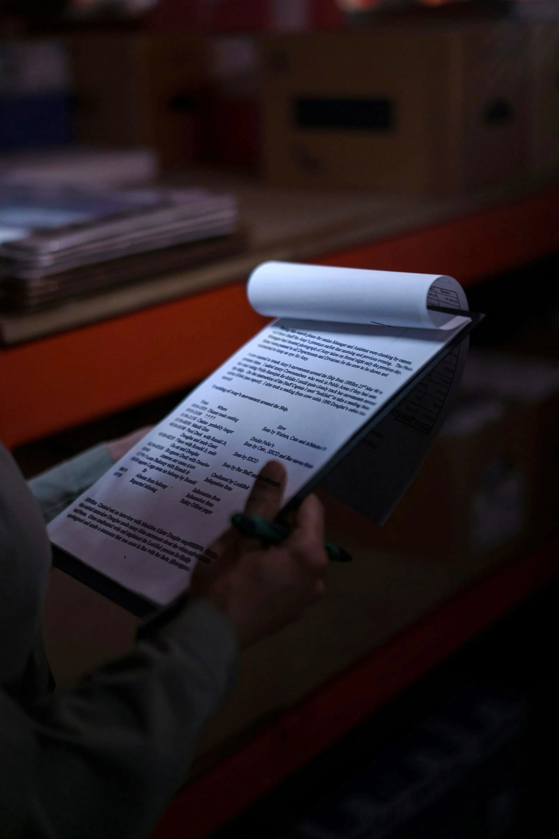 Person reading a printed receipt or document in a dimly lit room, with a table and stacked papers in the background.