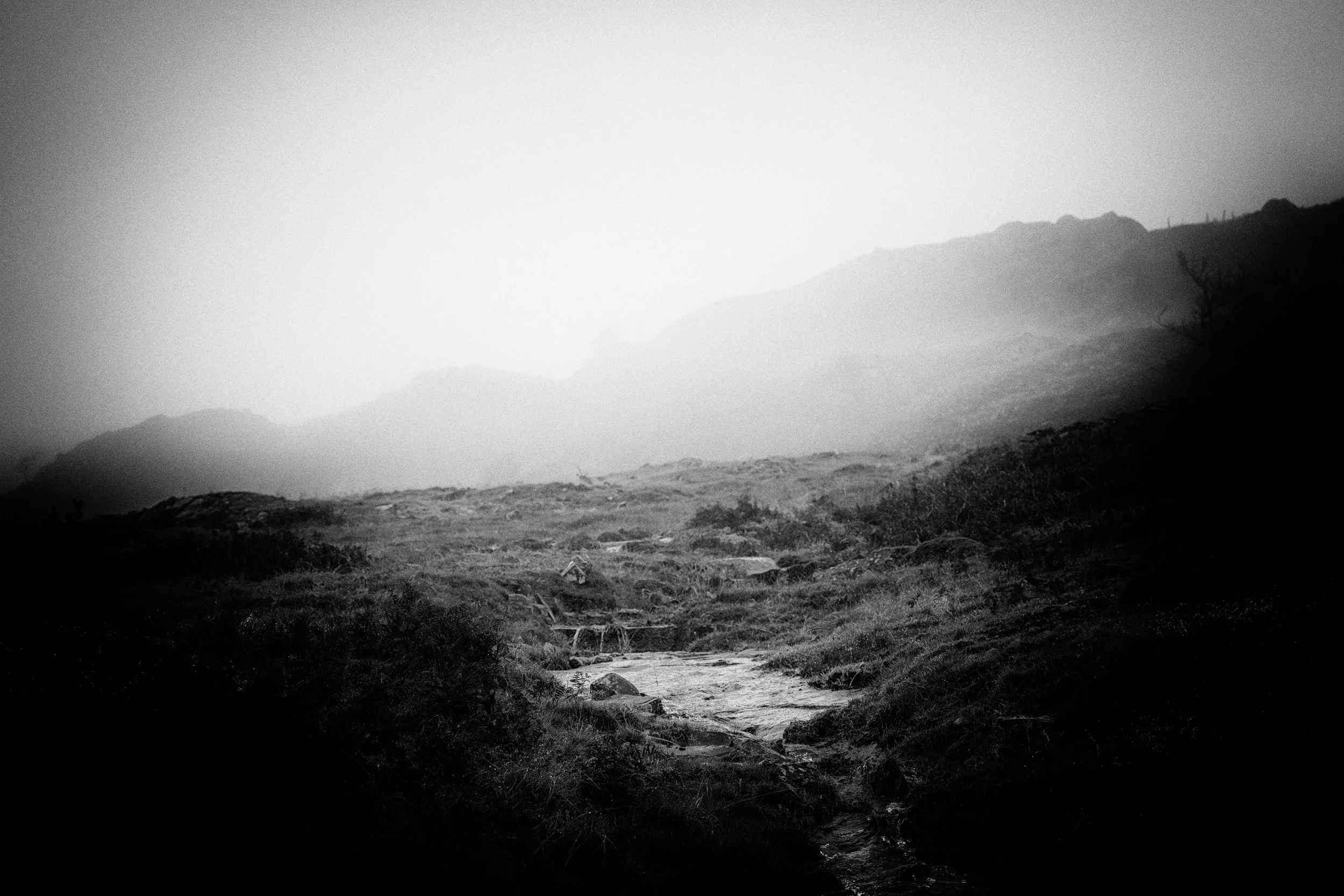 Black and white photo of a mountainous landscape with a small stream in the foreground and hills in the background.