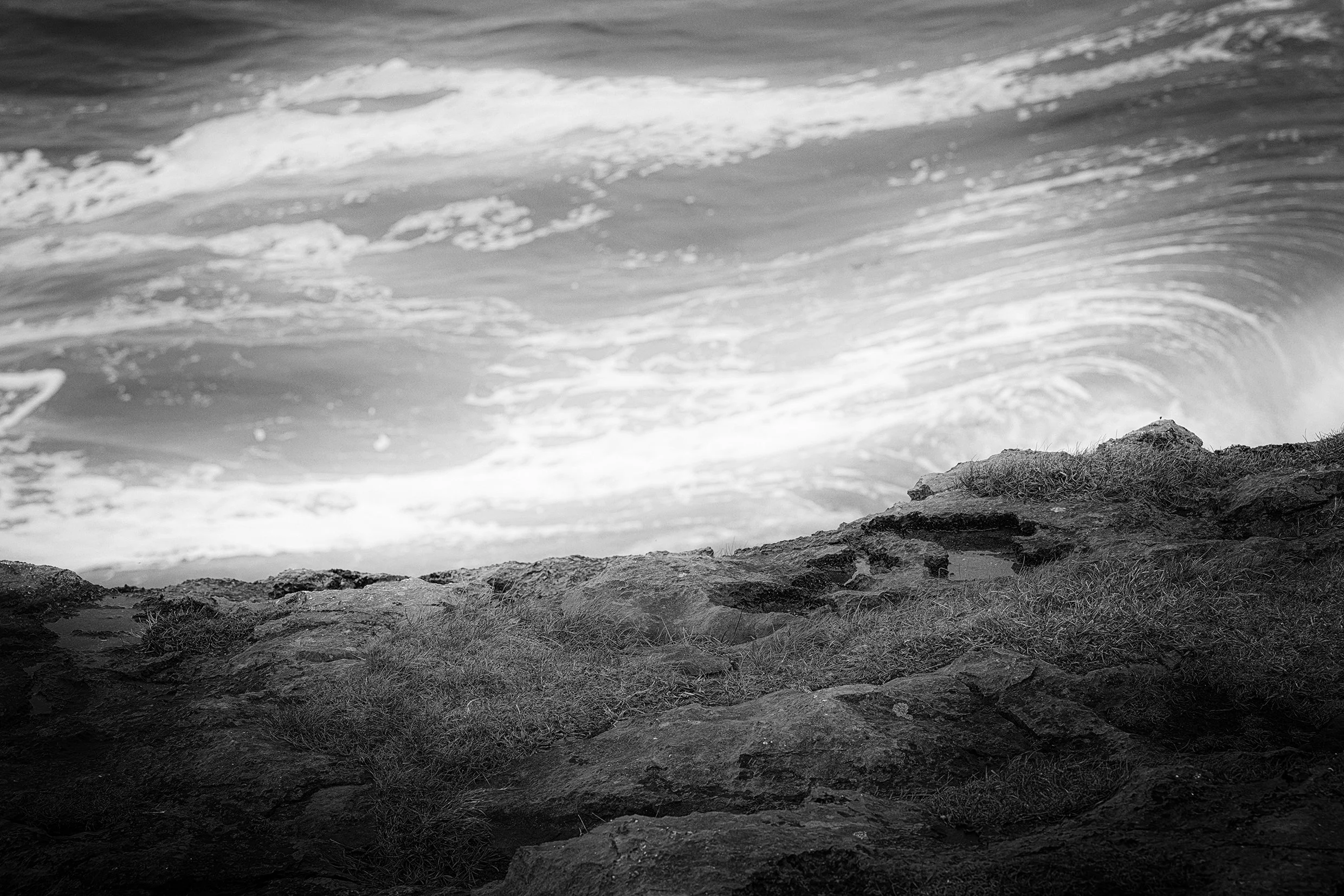 black and white negative landscape image showing a rough sea in the distance against the edge of a cliff