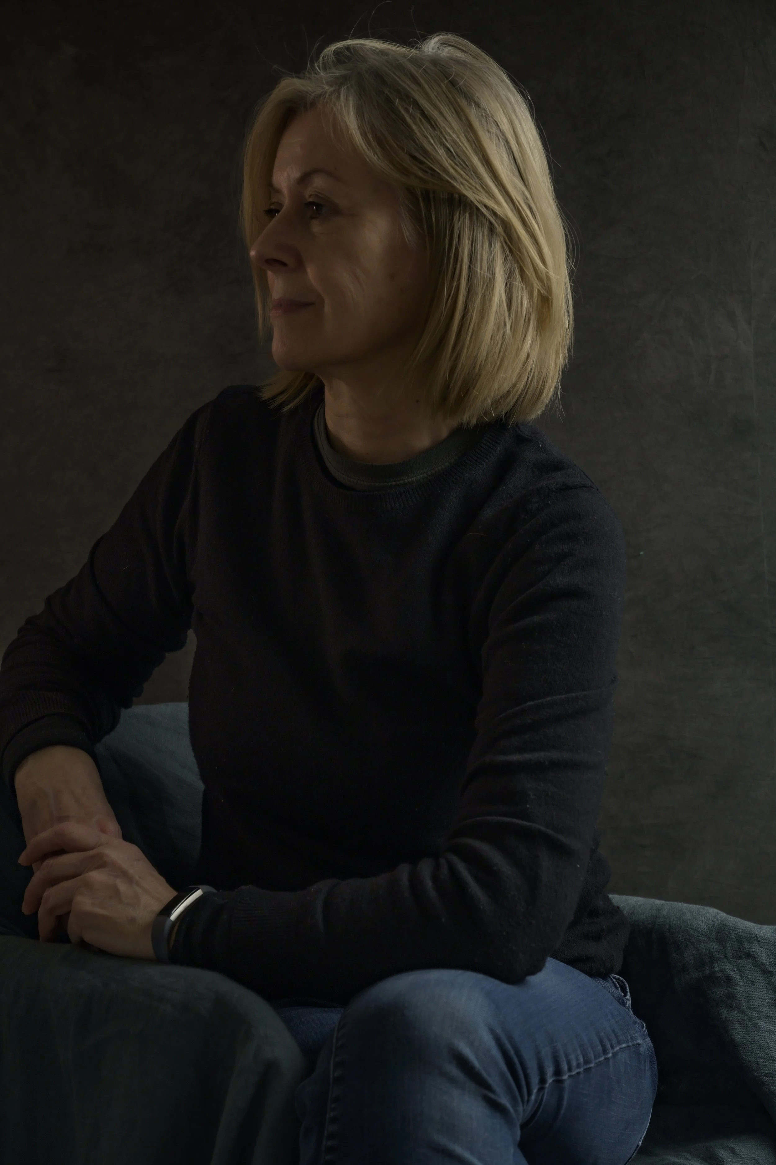 Side profile of a middle-aged woman with blonde hair, wearing a black sweater, sitting on a dark-colored armchair against a dark background.