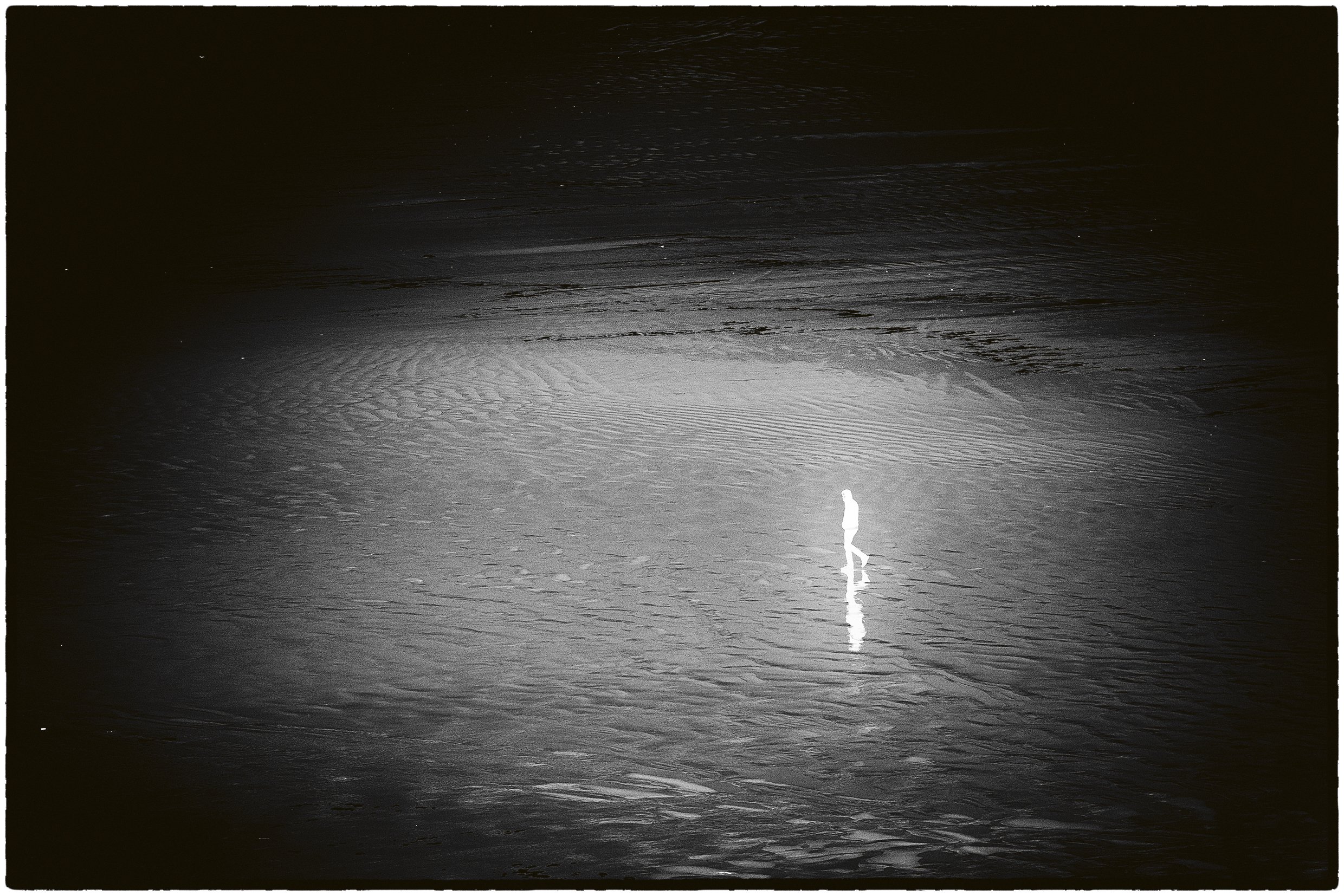 black and white negative landscape image showing a lone whited out figure walking in the black sand
