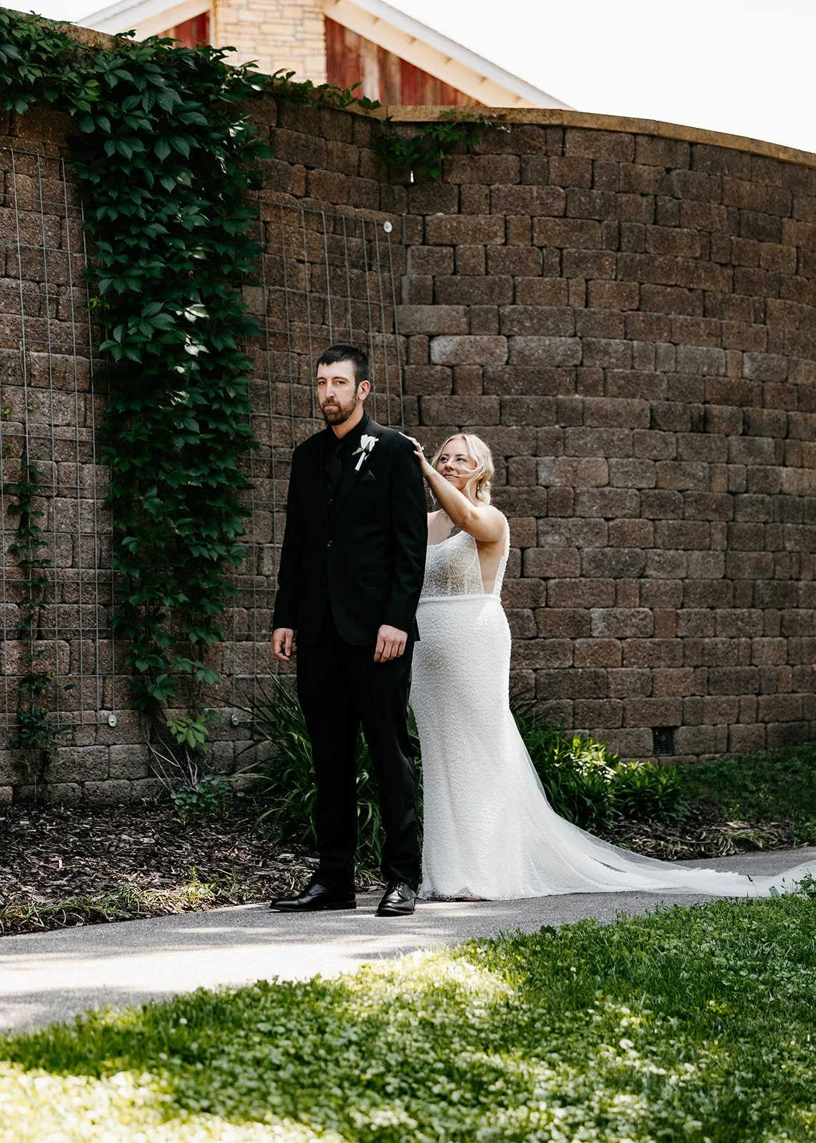 A bride in a white wedding dress is playfully touching the shoulder of a groom in a black suit, standing against a brick wall and greenery.