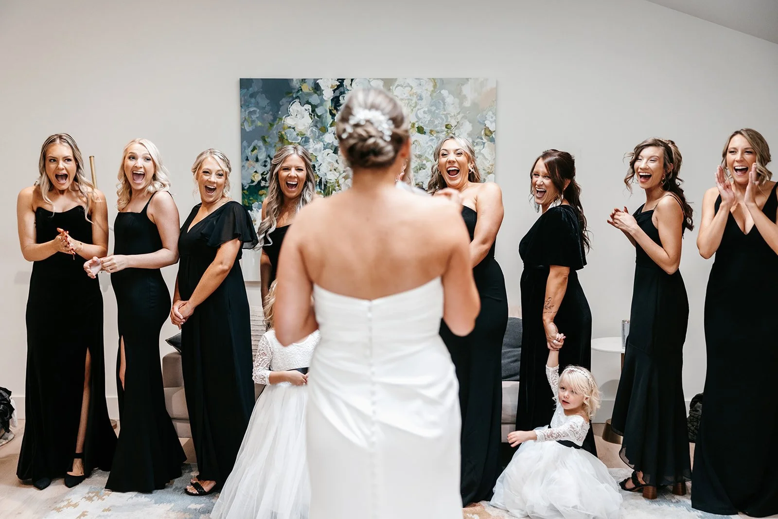 Bride holding a bouquet, seen from back, facing a group of smiling women in black dresses, with two young girls in white dresses in a room with white walls and a floral painting.
