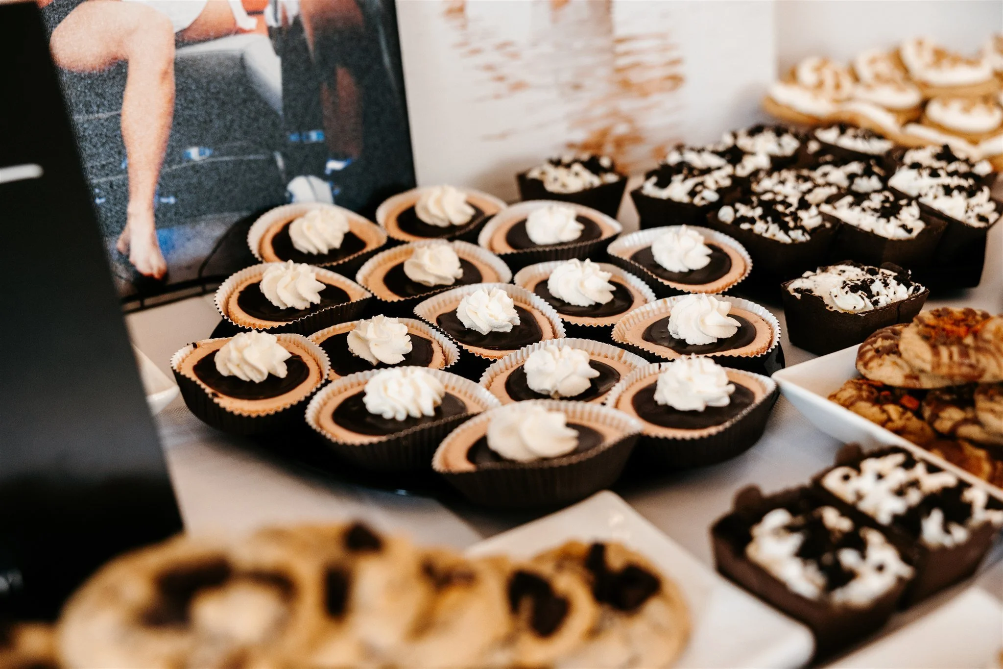 Assorted desserts including chocolate tarts with whipped cream, brownies topped with crushed cookies, and cookies on a platter, displayed on a table.