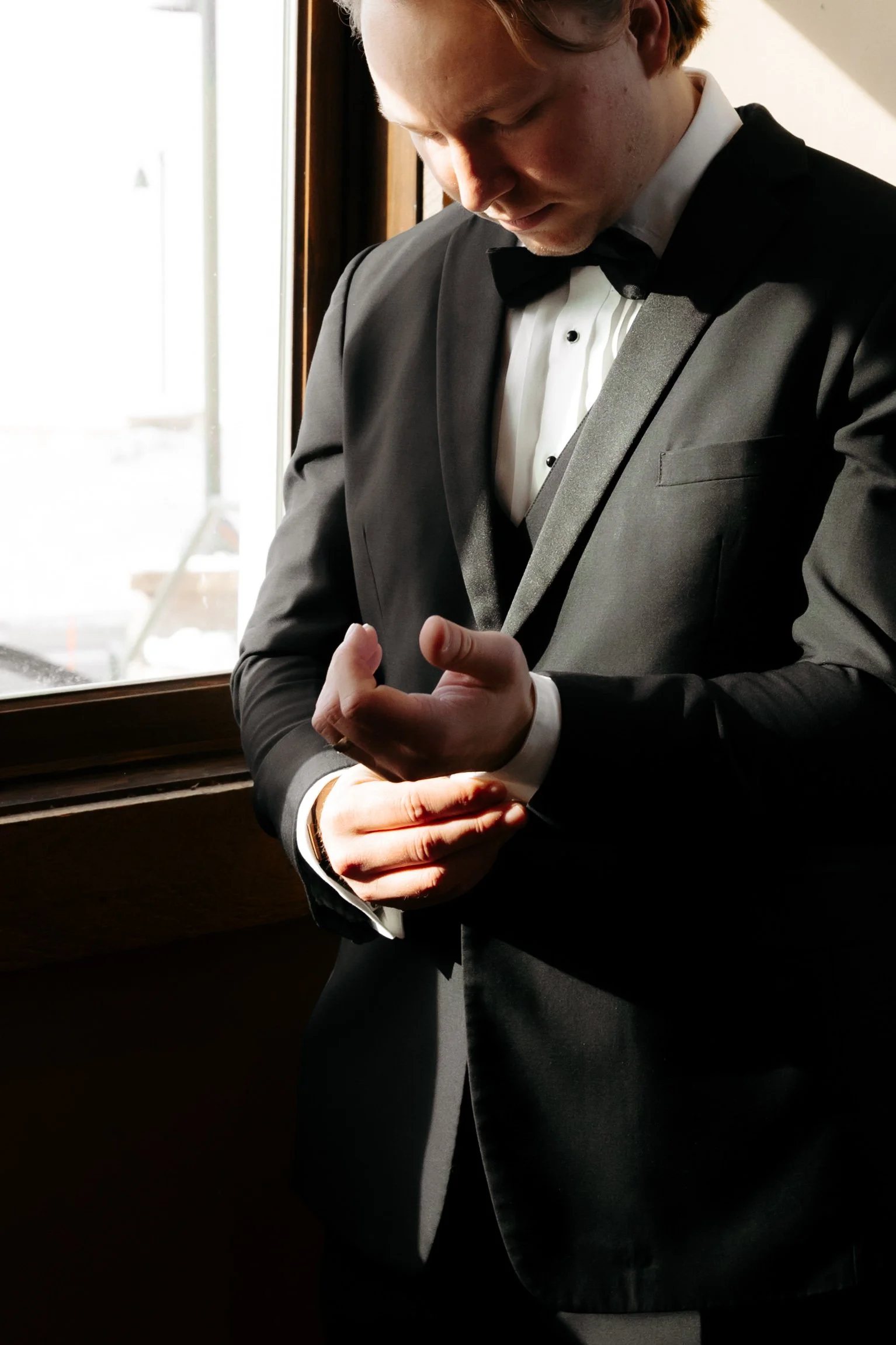 A man wearing a black tuxedo and bow tie standing by a window, adjusting his cufflinks in natural light.