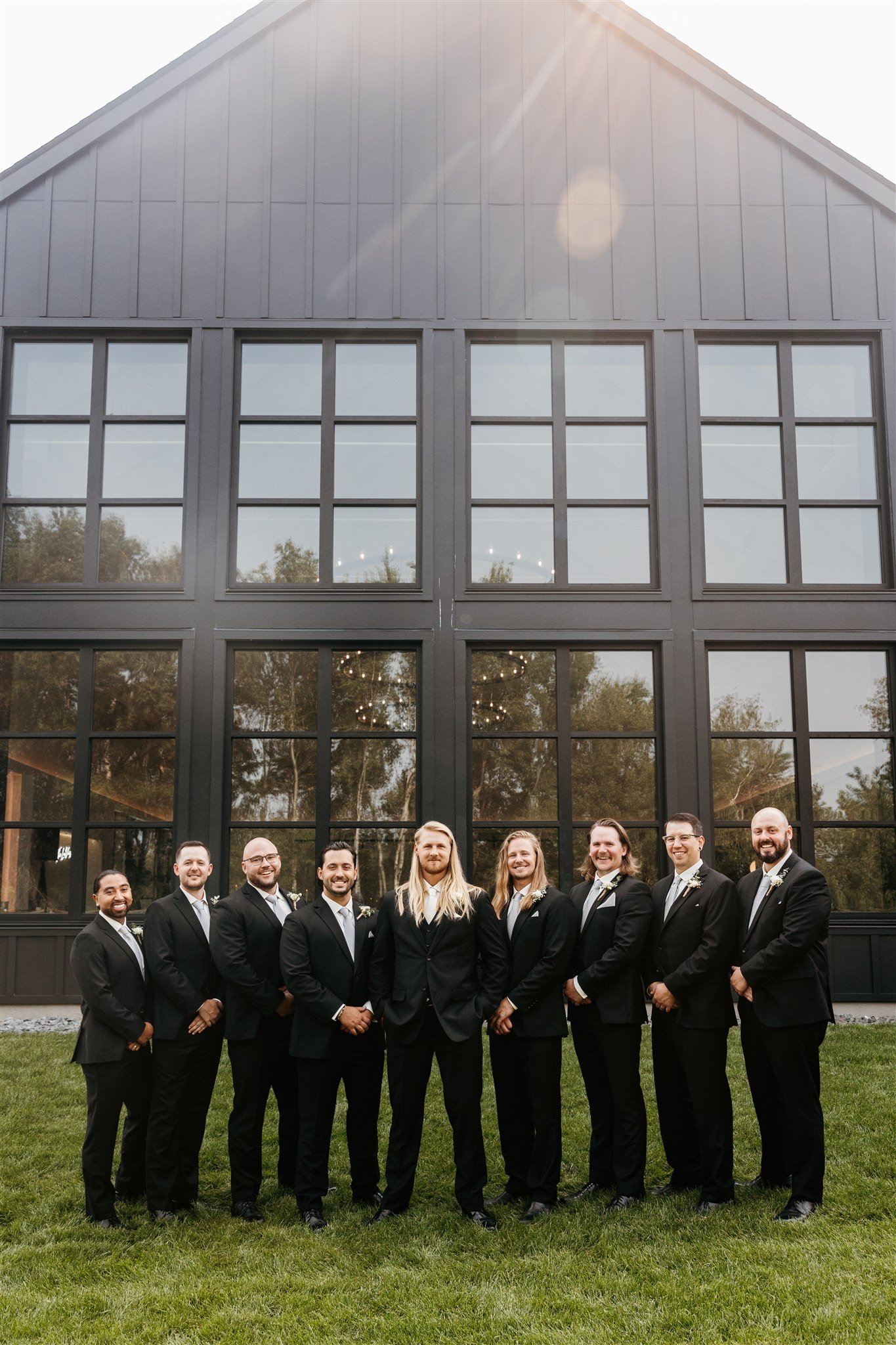 A group of nine men in formal suits standing outside in front of a large windowed building, smiling and posing for a photo.