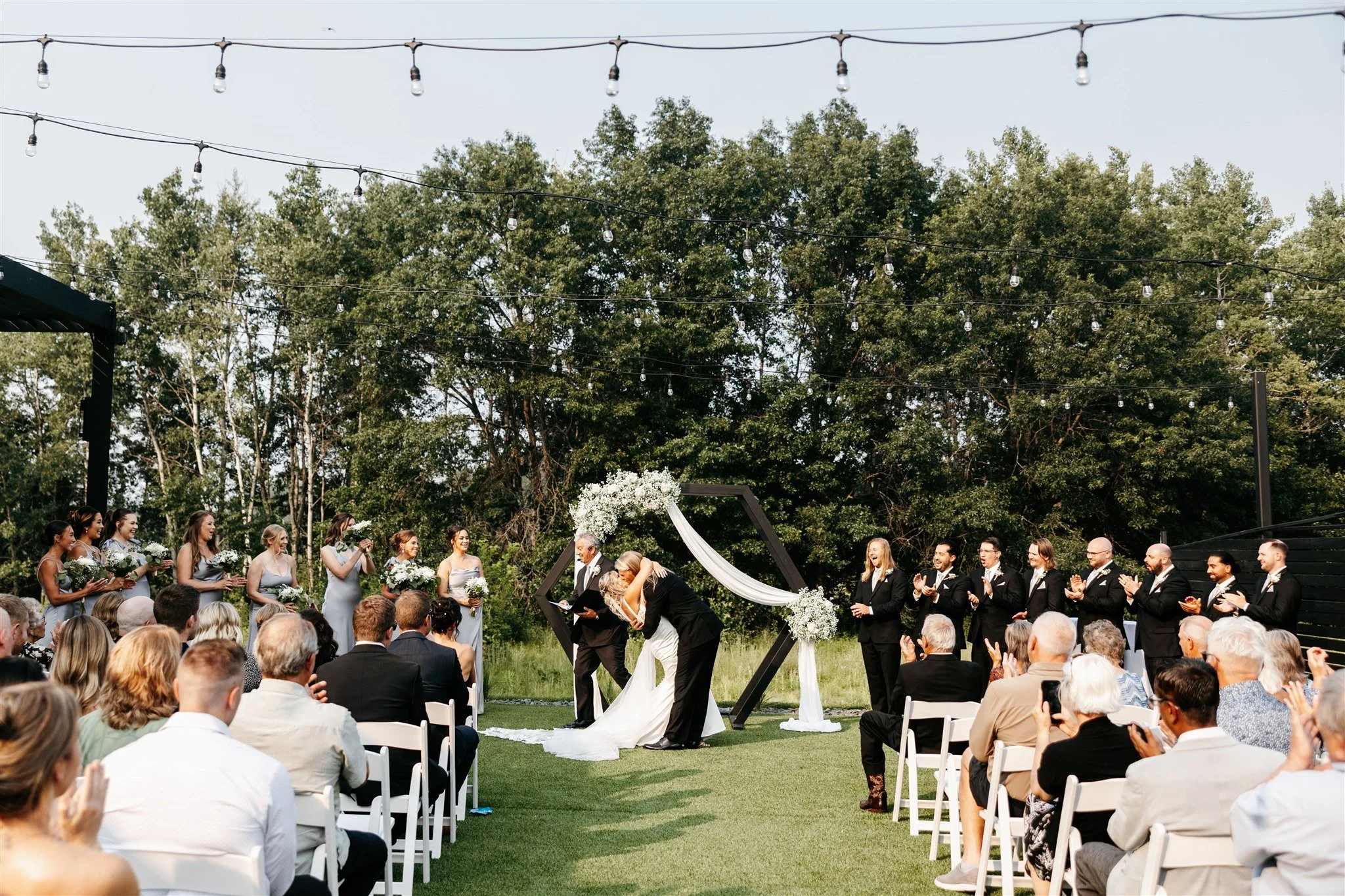 Outdoor wedding ceremony with a couple kissing, surrounded by bridesmaids and groomsmen, guests seated on white chairs, trees in the background, and string lights overhead.
