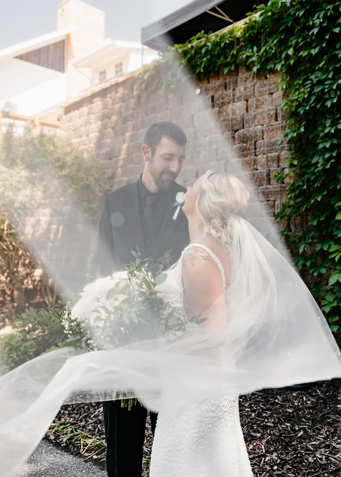 A bride and groom standing close to each other outdoors, with the bride holding a bouquet of white flowers and greenery. The bride is wearing a white wedding dress with beaded details and a veil, while the groom is dressed in a black suit. They are s