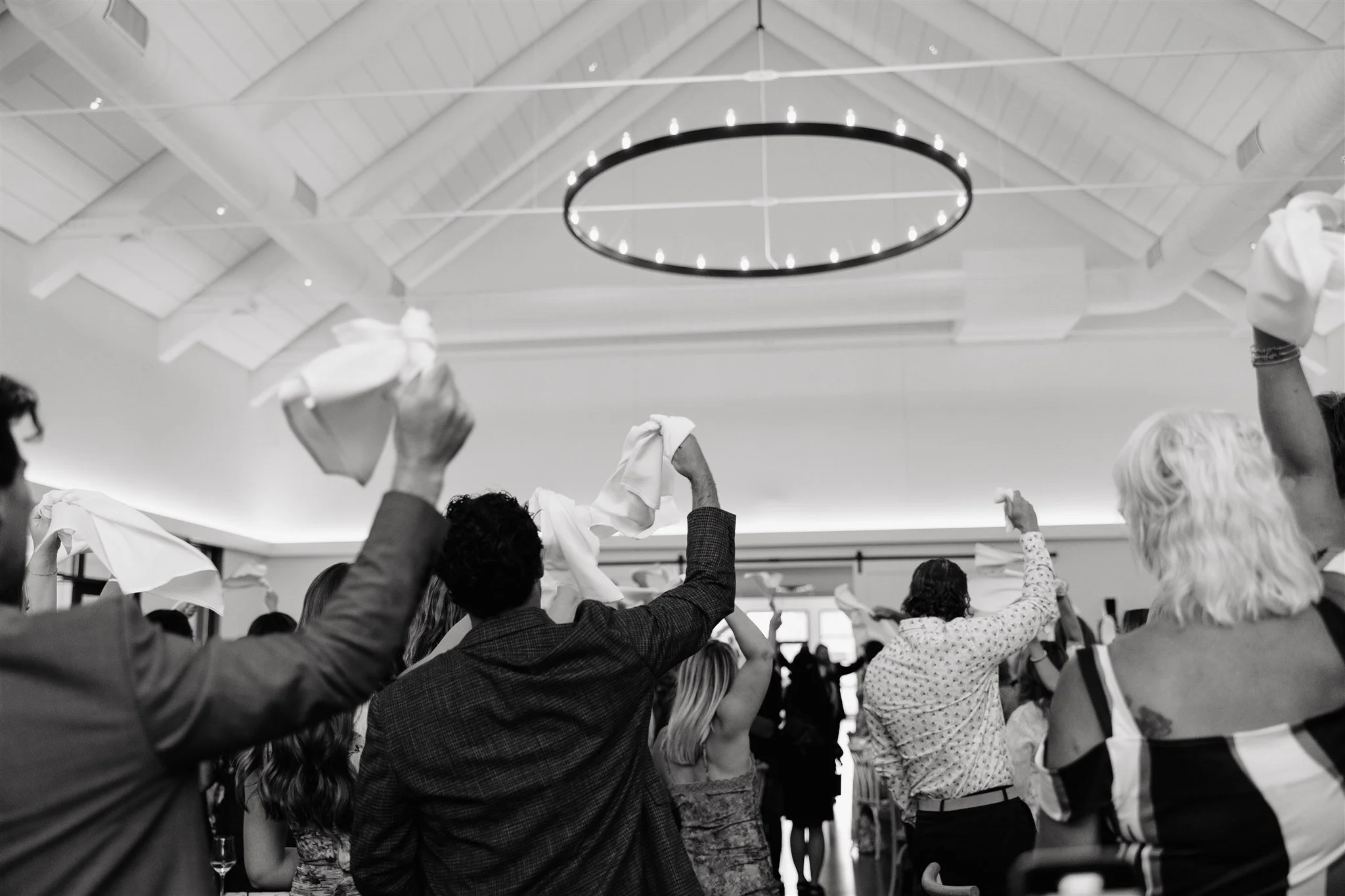 People raising cloth napkins in a celebration or gathering inside a room with a high ceiling and a circular lighting fixture.